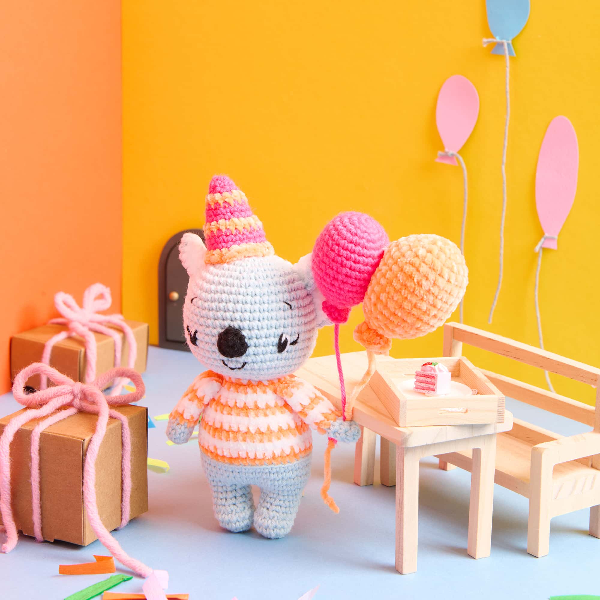 Ein gehäkelter Koala mit Partyhut hält Luftballons und steht in einer bunt dekorierten Szene mit Partydekoration.