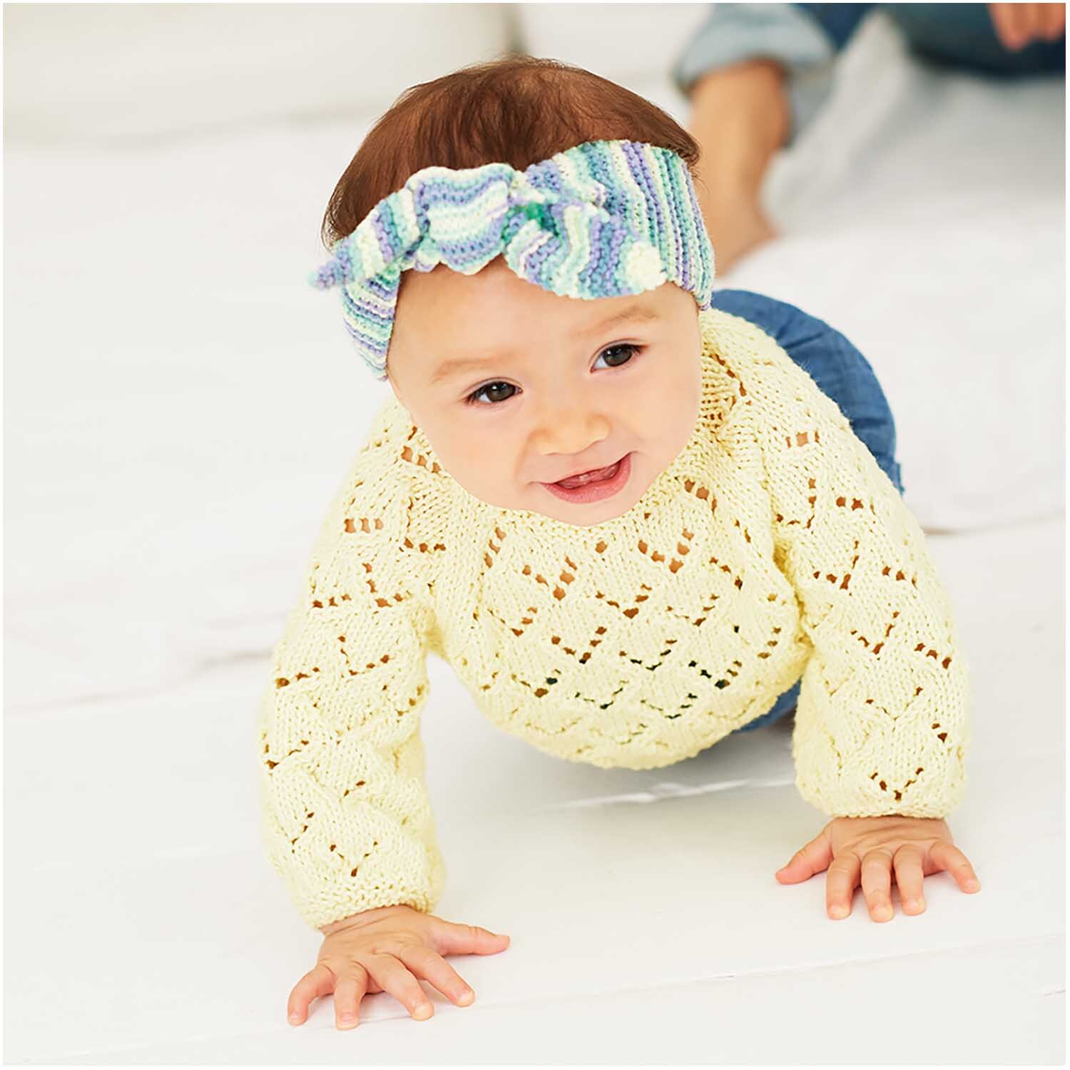 Rico Baby Cotton Soft dk: Ein Baby in einem gestrickten pastellgelben Pullover krabbelt auf einer weißen Decke.