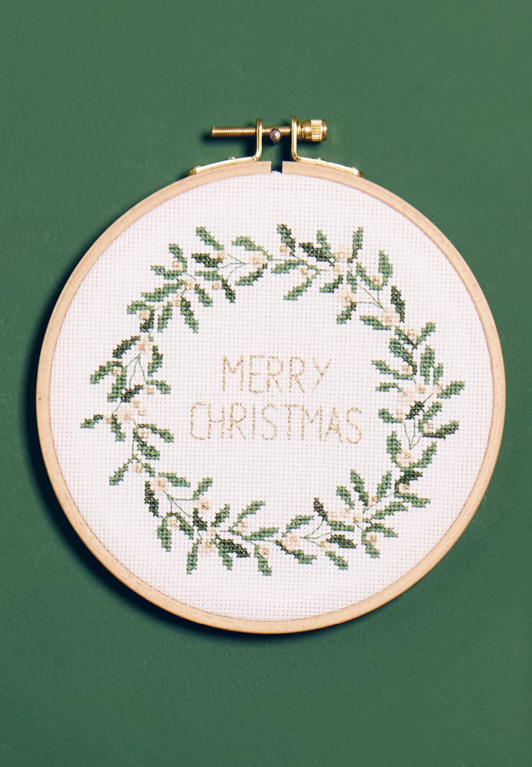 Ein Stickbild mit Weihnachtskranz und dem Schriftzug "Merry Christmas" hängt an einer dunkelgrünen Wand. 