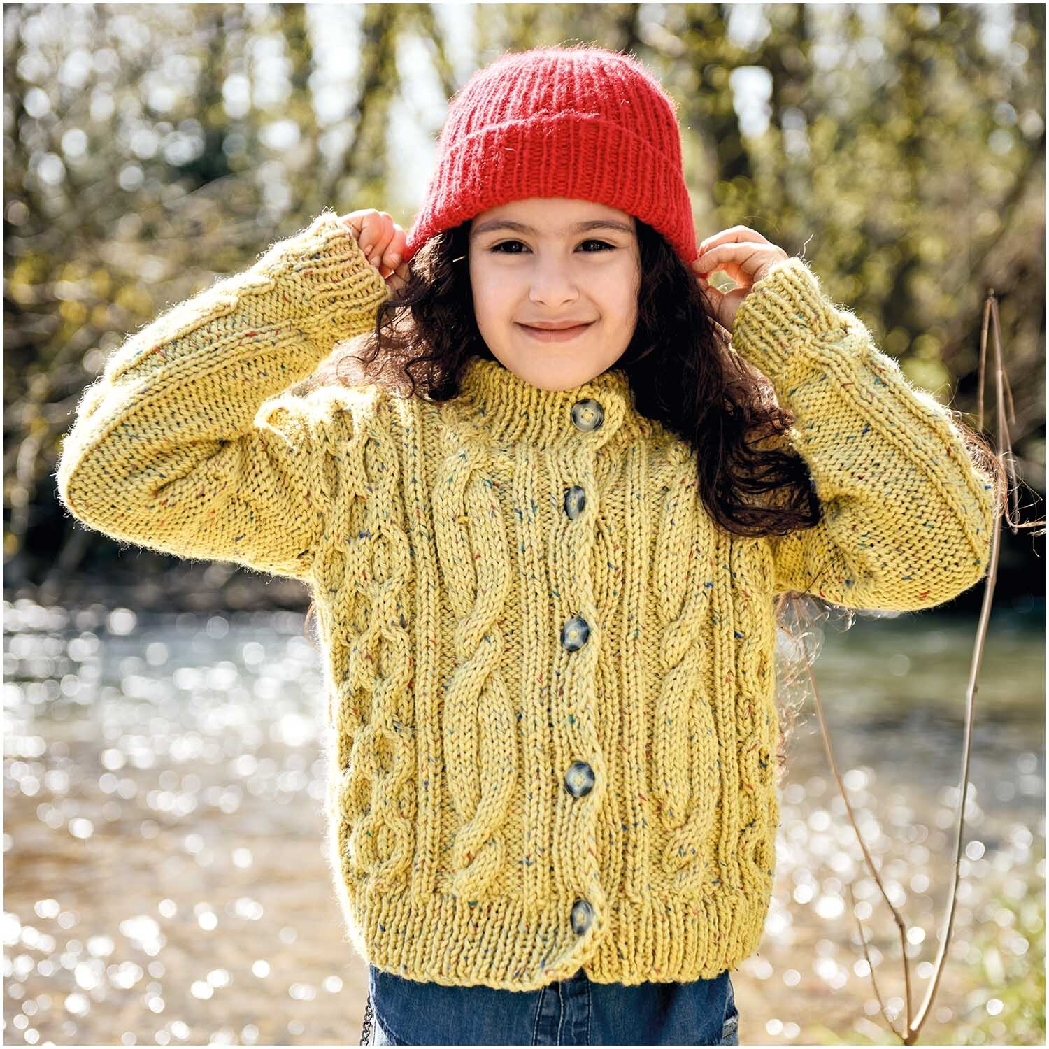 Essentials Mega Wool Tweed chunky: Kind in gelbem Strickpullover und roter Mütze von Rico Design am Flussufer.