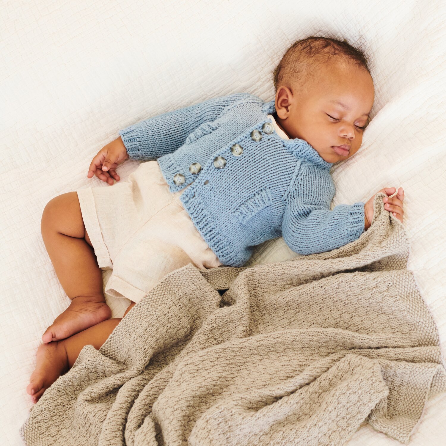 Strickset Jacke Modell 15 aus Baby 043: Schlafendes Baby in blauer Strickjacke auf beiger Decke.