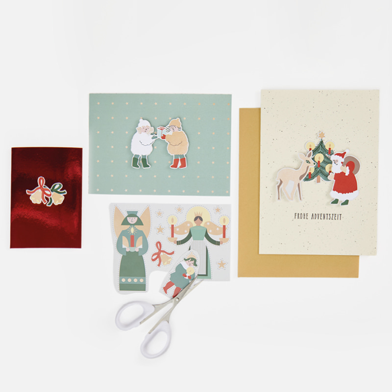 Karten mit weihnachtlichen Motiven liegen umgeben von Bastelmaterial auf hellgrauem Hintergrund