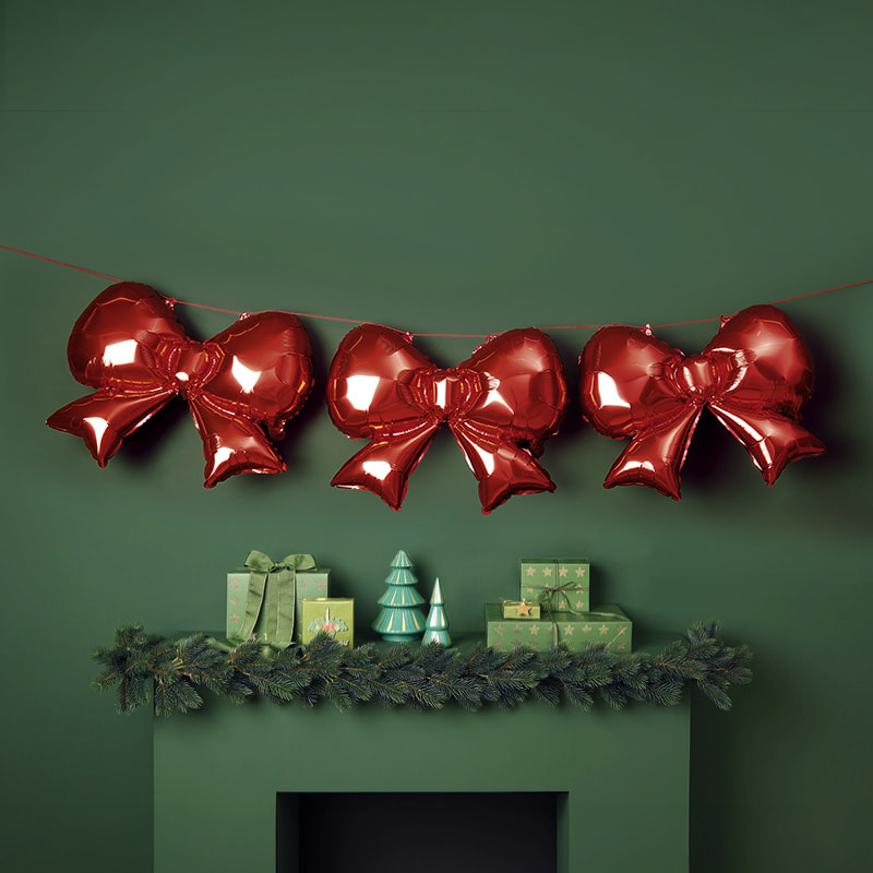 Eine Girlande mit Folienballons in Schleifenform hängt dekorativ vor einem weihnachtlich geschmückten Regal. 