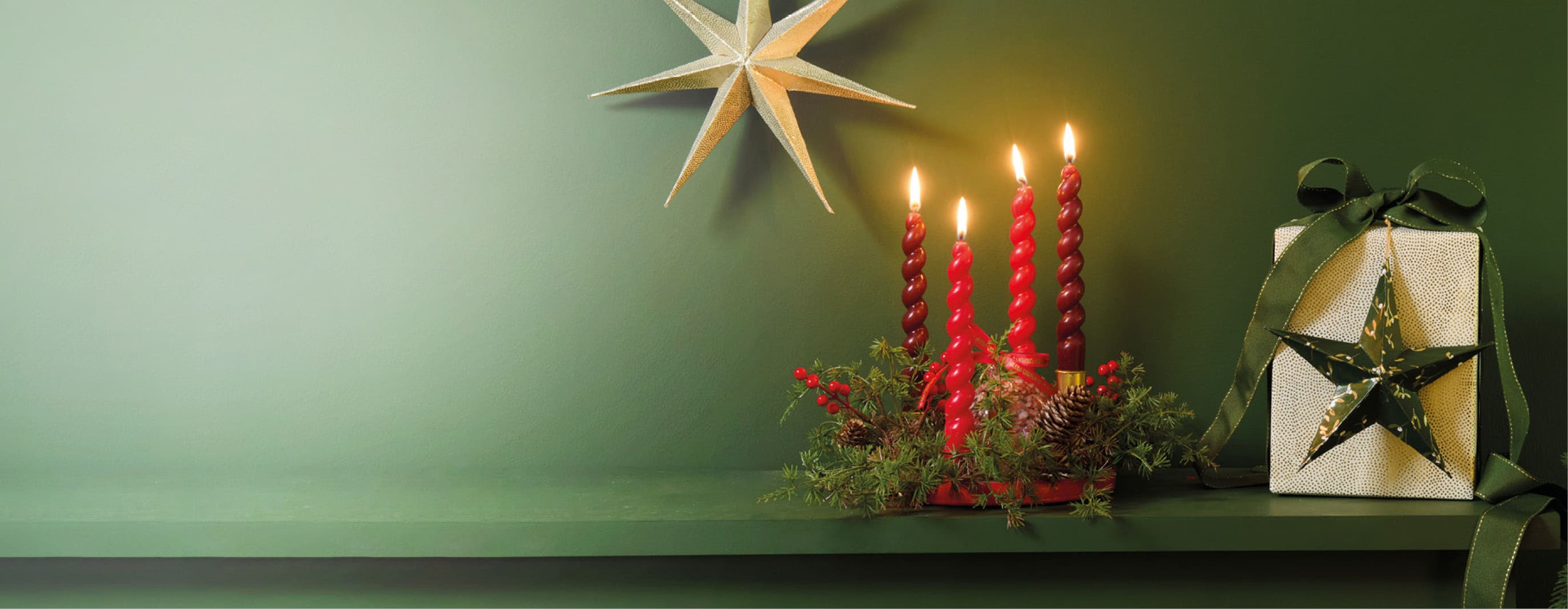 Moderner Adventskranz mit vier roten Kerzen, Tannengrün und roten Beeren vor grüner Wand. Daneben stehen ein Geschenk und ein leuchtender Papierstern.