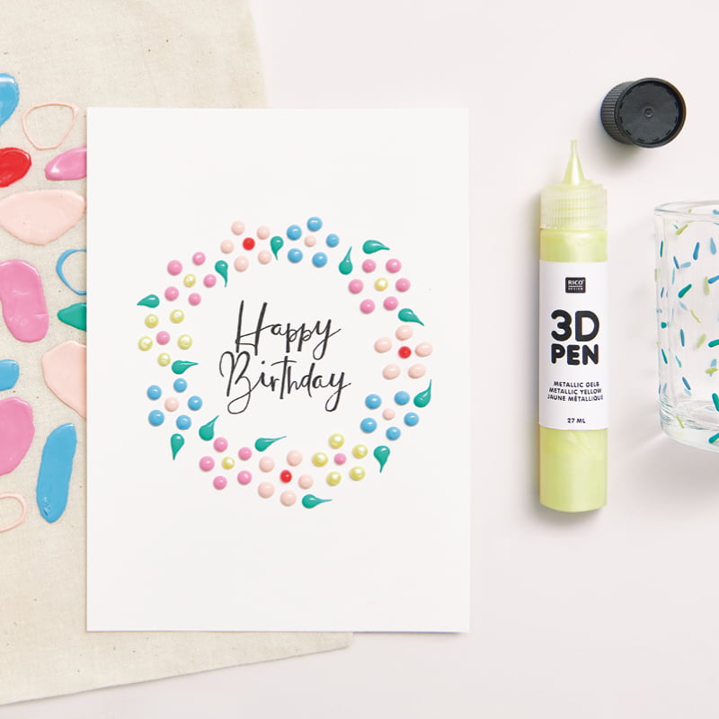Bemalte Happy Birthday-Karte mit 3D Punkten liegt zusammen mit einem 3D Pen auf grauem Hintergrund.