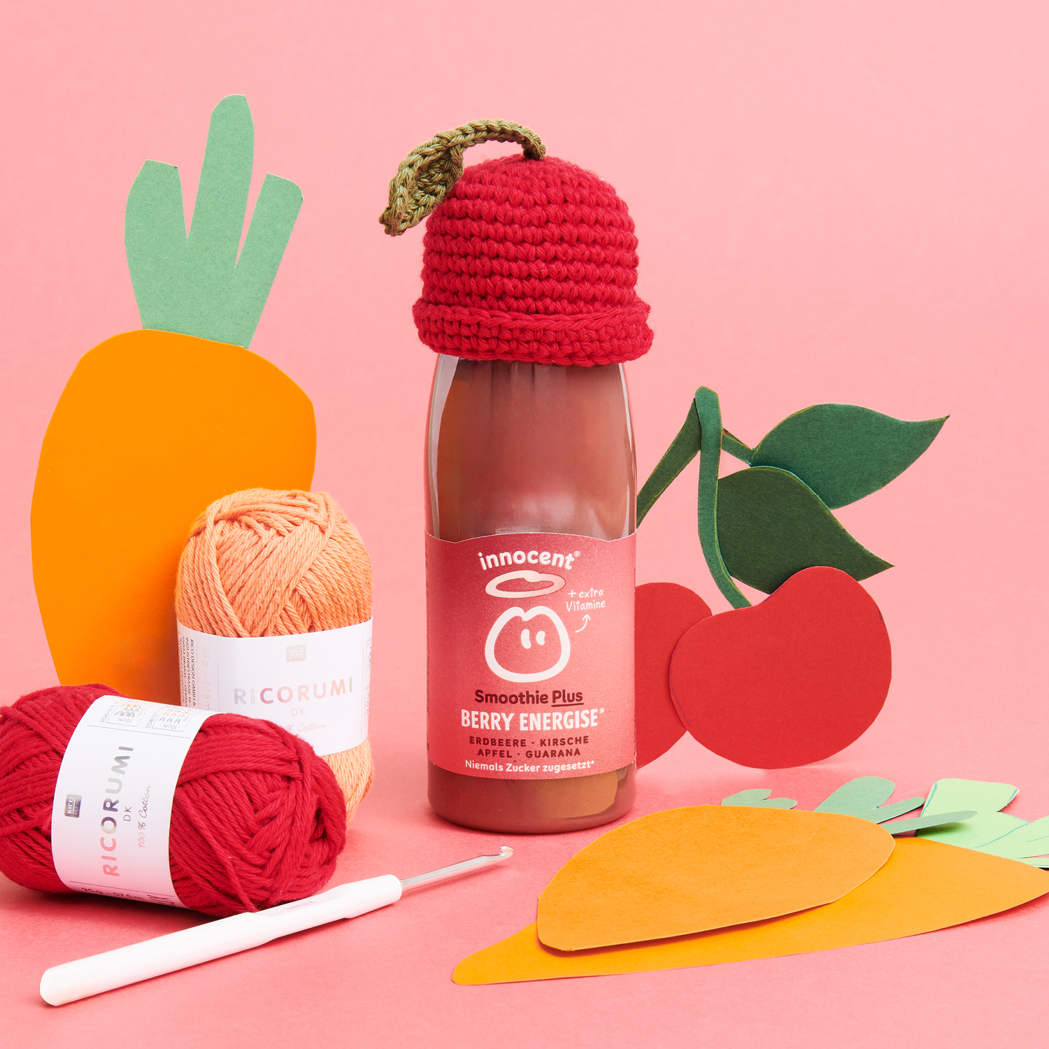 Häkelanleitung Ricorumi Mützchen Kirsche: Rotes Mützchen auf Smoothie-Flasche, umgeben von Garn und Papierdekorationen.