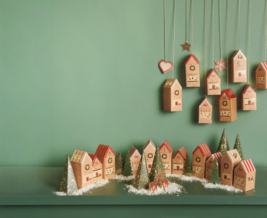 24 Häuser und Tannenbäume aus Papier in einem weihnachtlichen Design stehen dekoriert vor einer grünen Wand.