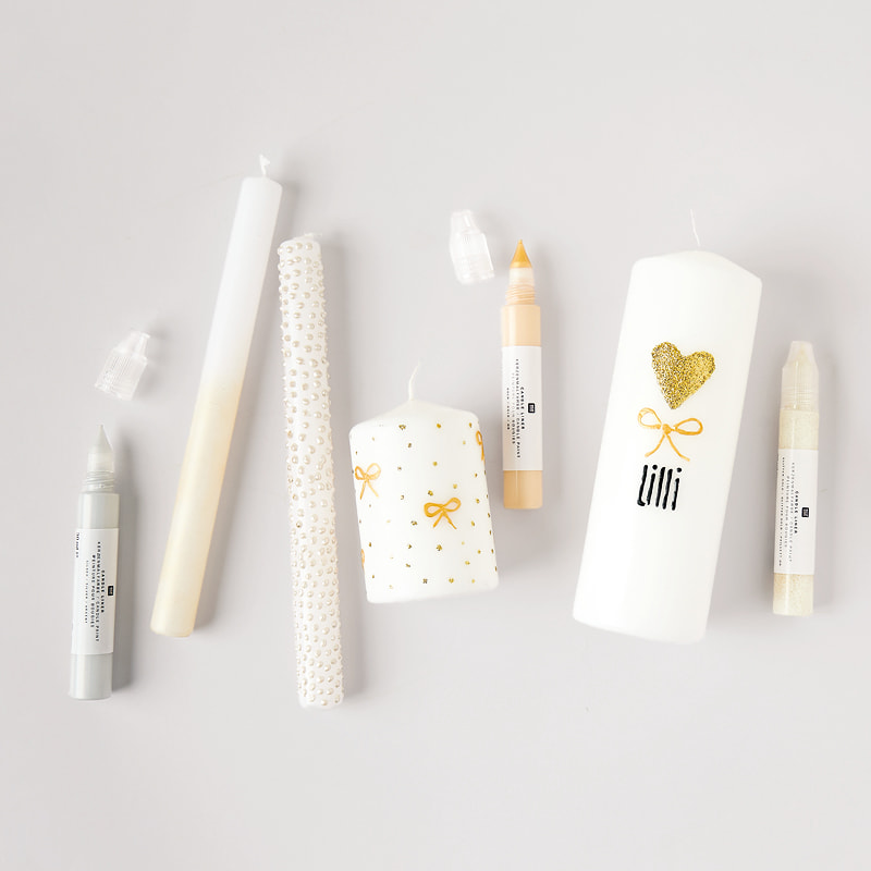 Kerzen mit goldenen und silbernen Verzierungen liegen zusammen mit Kerzenmalstiften auf grauem Hintergrund.