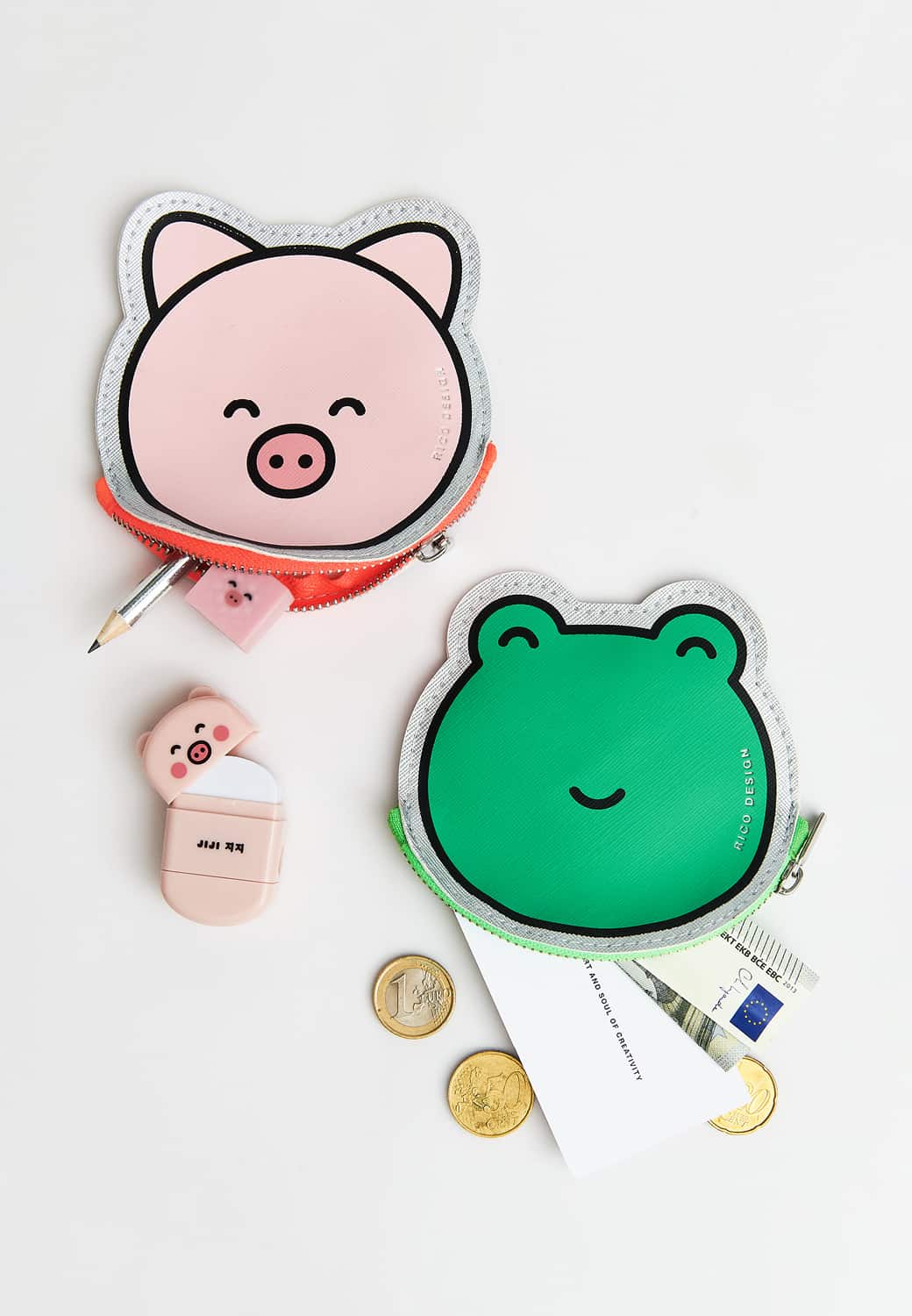 Zwei Portmonnaies in Form von Frosch und Schwein liegen umgeben von Geld und Kleinigkeiten auf grauem Hintergrund.