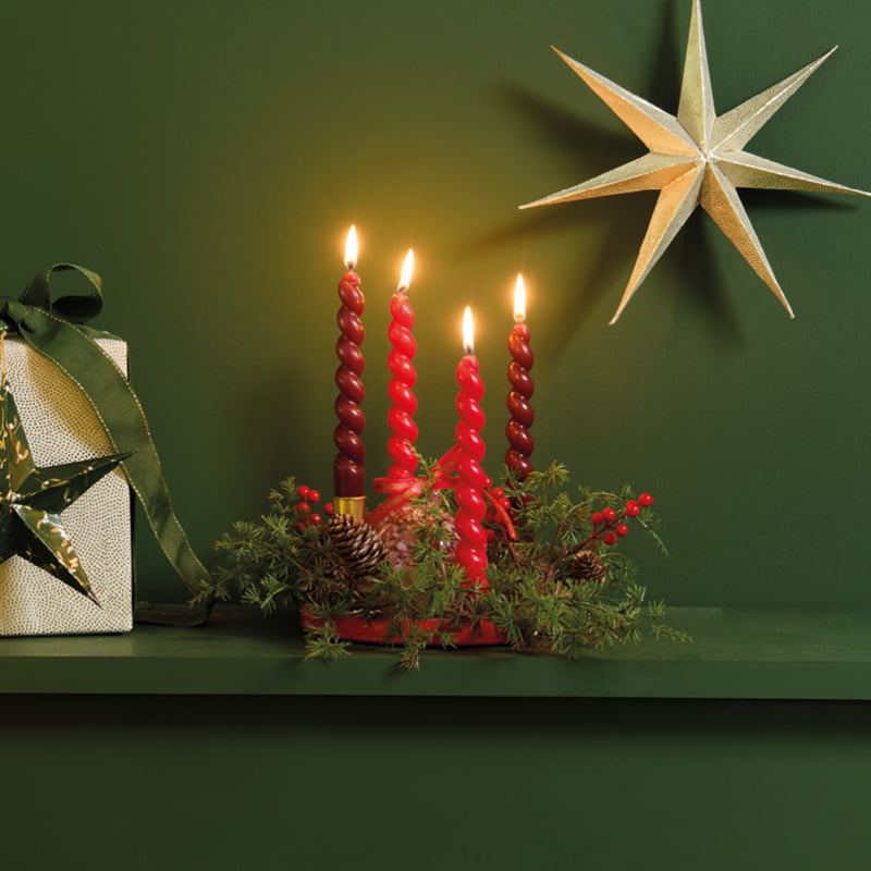 Ein Adventskranz ist vor einer dunkelgrünen Wand mit Tannenzweigen, Beeren und roten Kerzen dekoriert.