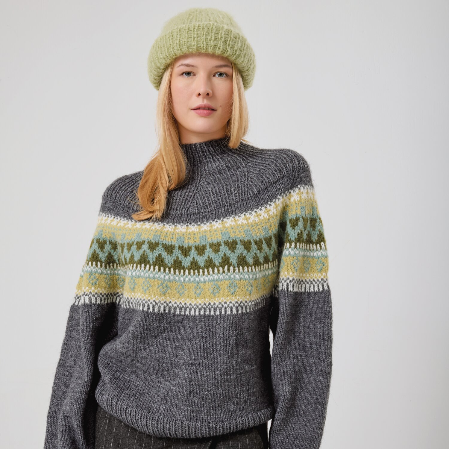 Strickset Pullover Modell 01 aus Made by Me No. 21 von Rico Design Strickset Pullover Modell 01 aus Made by Me No. 21: Grauer Pullover mit buntem Muster, getragen von Frau mit grüner Mütze.