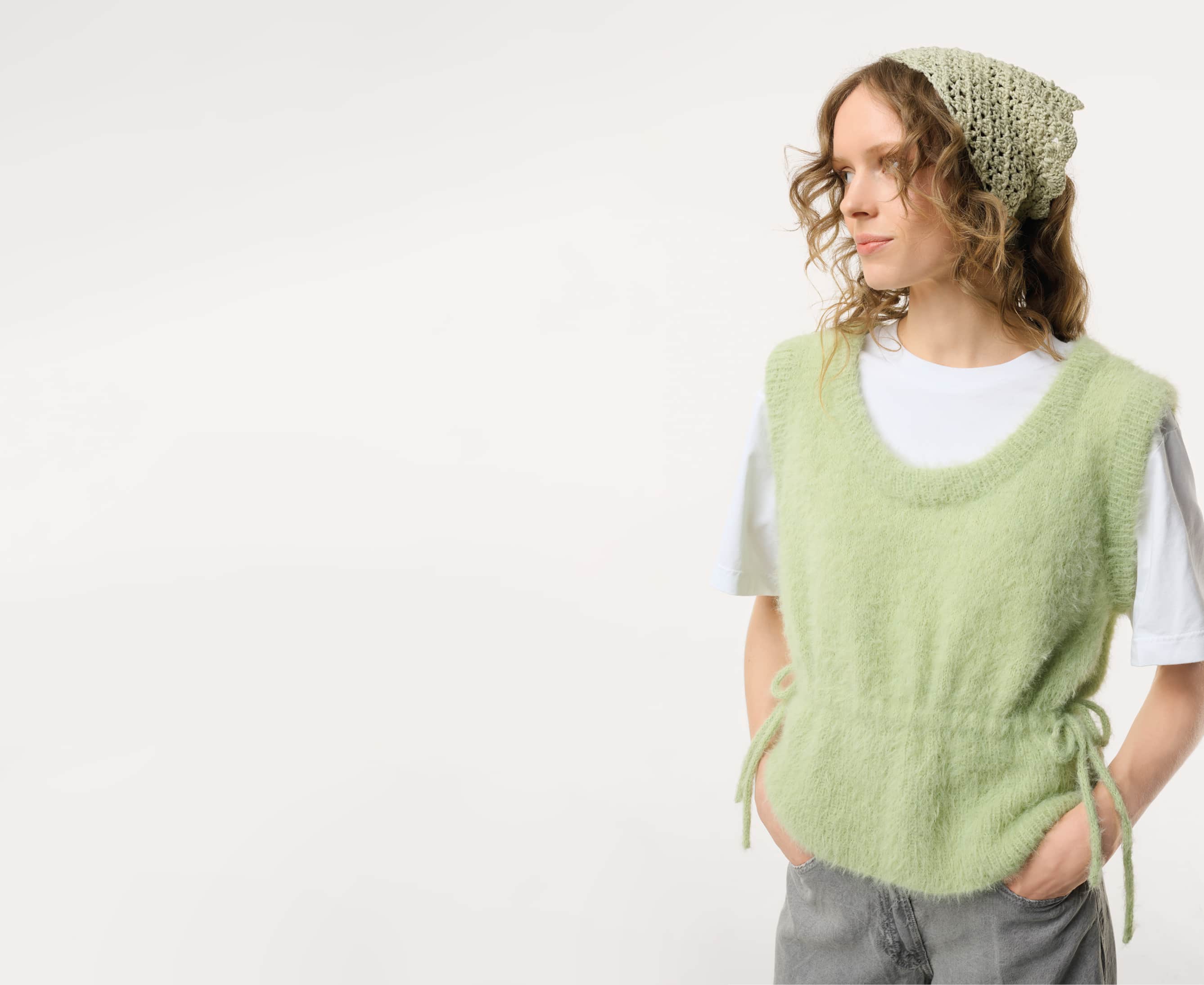 Eine Frau trägt eine gehäkeltes Kopftuch und einen gestrickten Pullunder und posiert vor grauem Hintergrund.