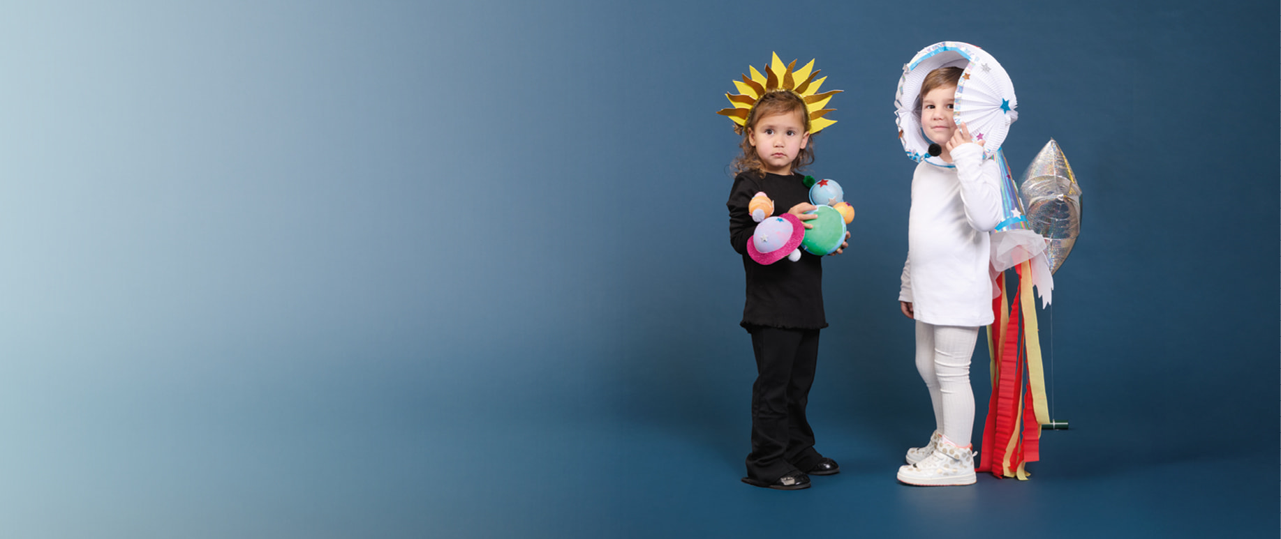 Zwei Kinder stehen in Karnevalskostümen vor dunkelblauem Hintergrund. Ein Kind trägt eine Sonnenkrone und eine Planetenkette. Das andere Kind hat ein Astronauten-Kostüm an. 