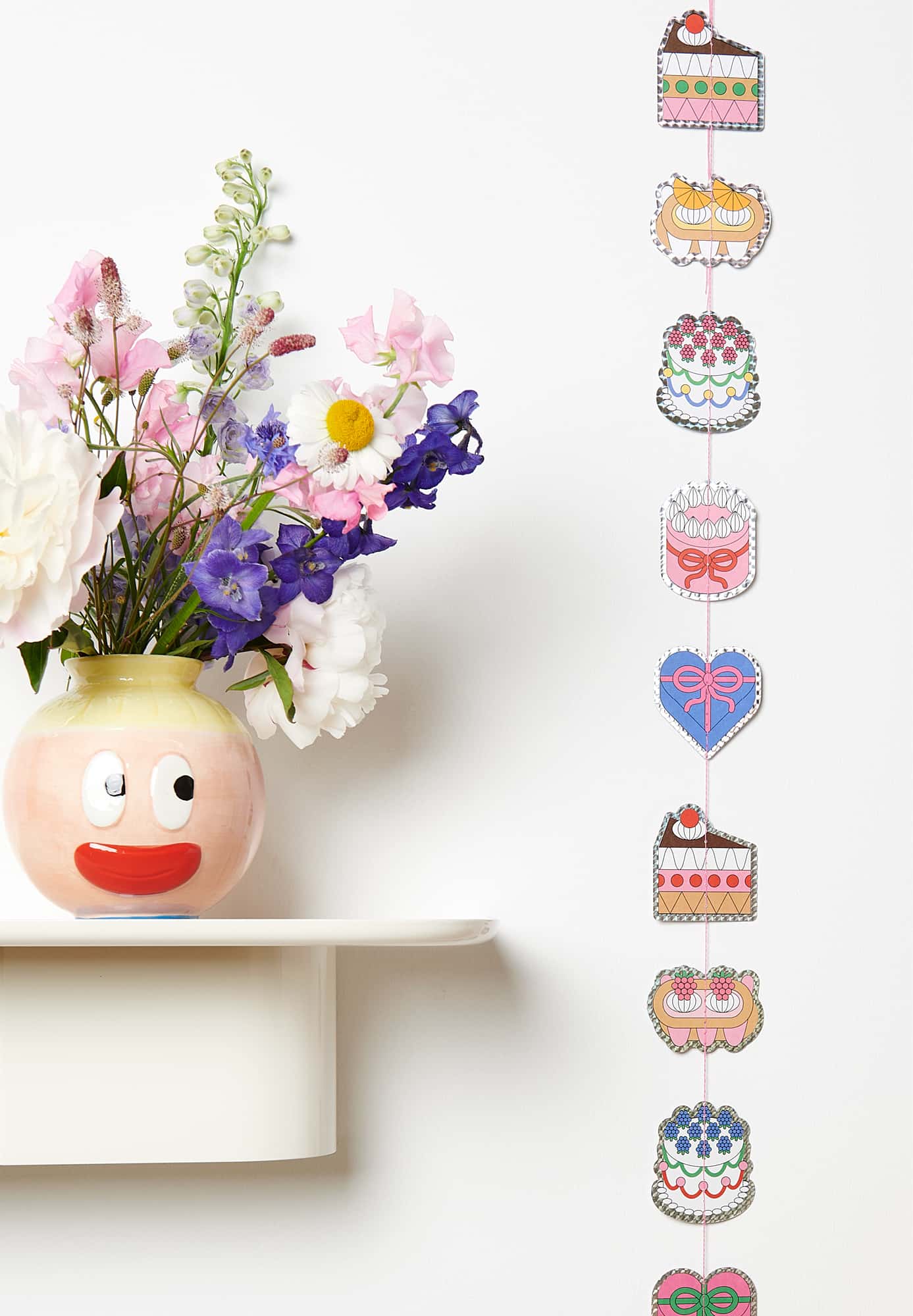 Eine Girlande mit illustrierten Torten hängt an einer Wand. Daneben eine mit Blumen gefüllte Vase mit Gesicht. 