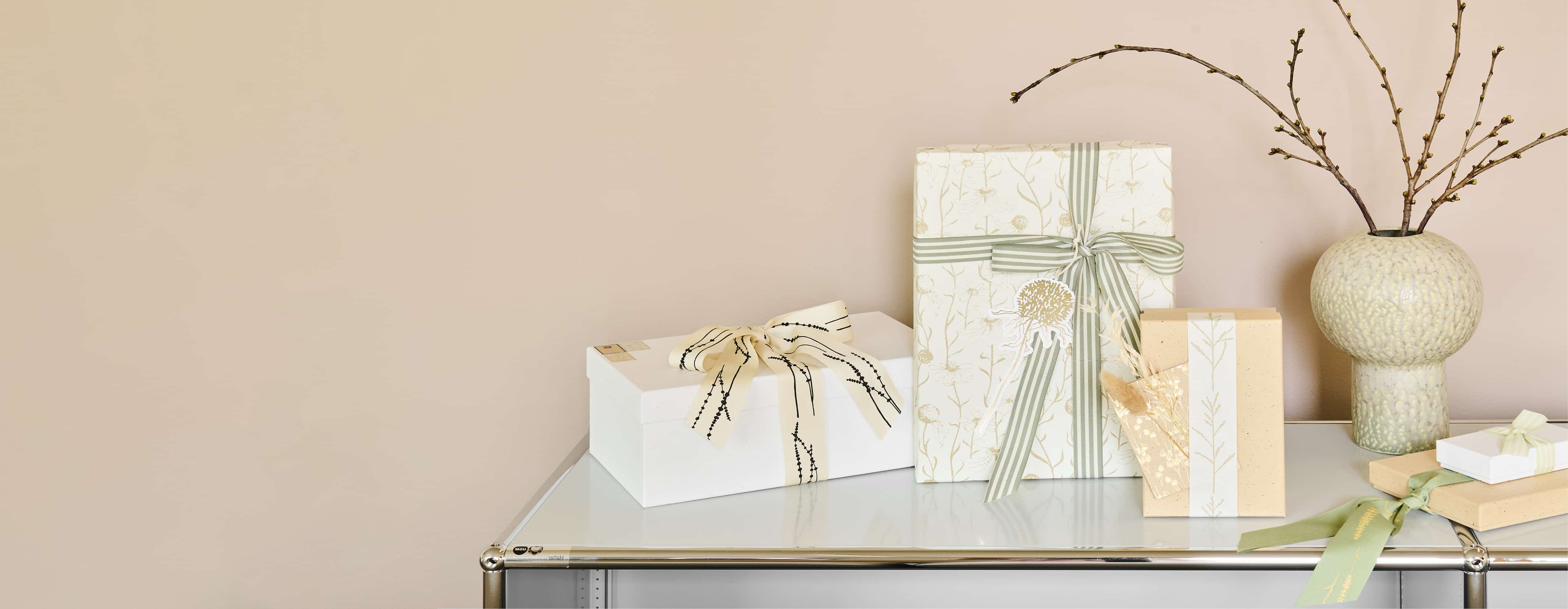 Verpackte Geschenke in Naturtönen und mit Details von Gräsermotiven stehen dekorativ auf einem weißen Regal. 