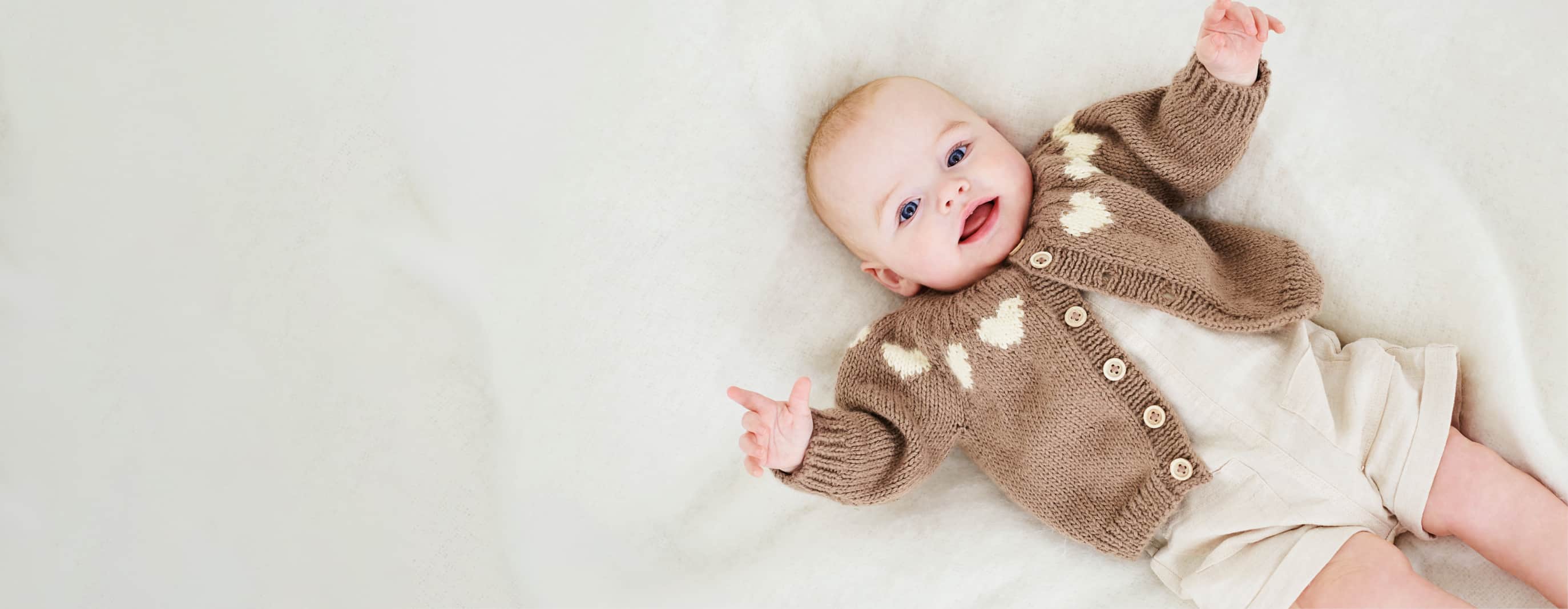 Ein Baby liegt mit einer Strickjacke gekleidet auf einer Decke. 