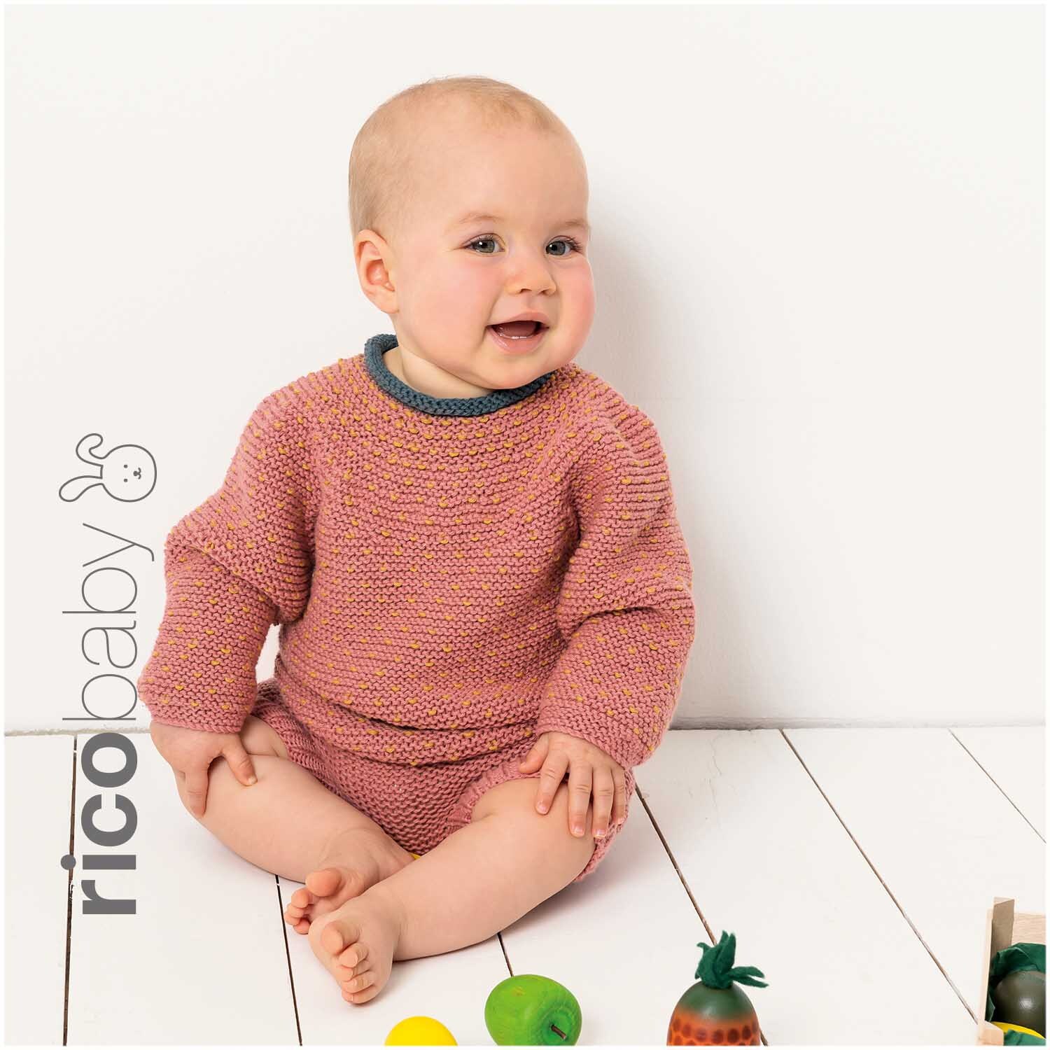 Rico Baby Cotton Soft dk: Ein Baby trägt einen gestrickten Pullover und Shorts, umgeben von Spielzeug.