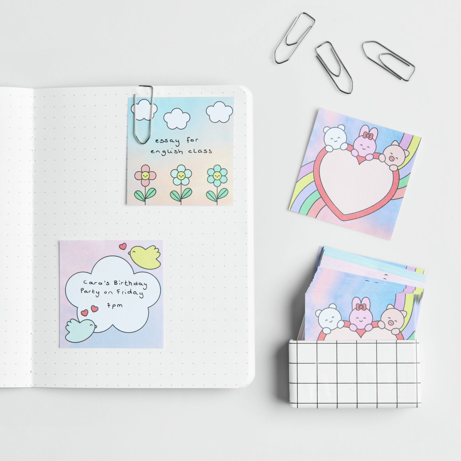 Paper Poetry Mini-Notizzettel-Box Chingu mit bunten Designs, 5x5cm, 120 Blatt, neben einem Notizbuch und Büroklammern.