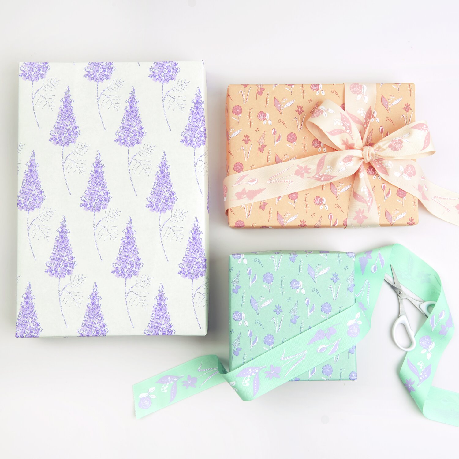 Geschenkpapier Blumenwiese: Drei Geschenke in floralen Designs, verpackt in grünem und orangem Papier, mit Schleifen und Schere.