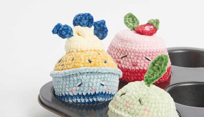 Häkelanleitung Blueberry Cupcake: Drei bunte, gehäkelte Cupcakes in einer Muffinform, verziert mit Früchten.