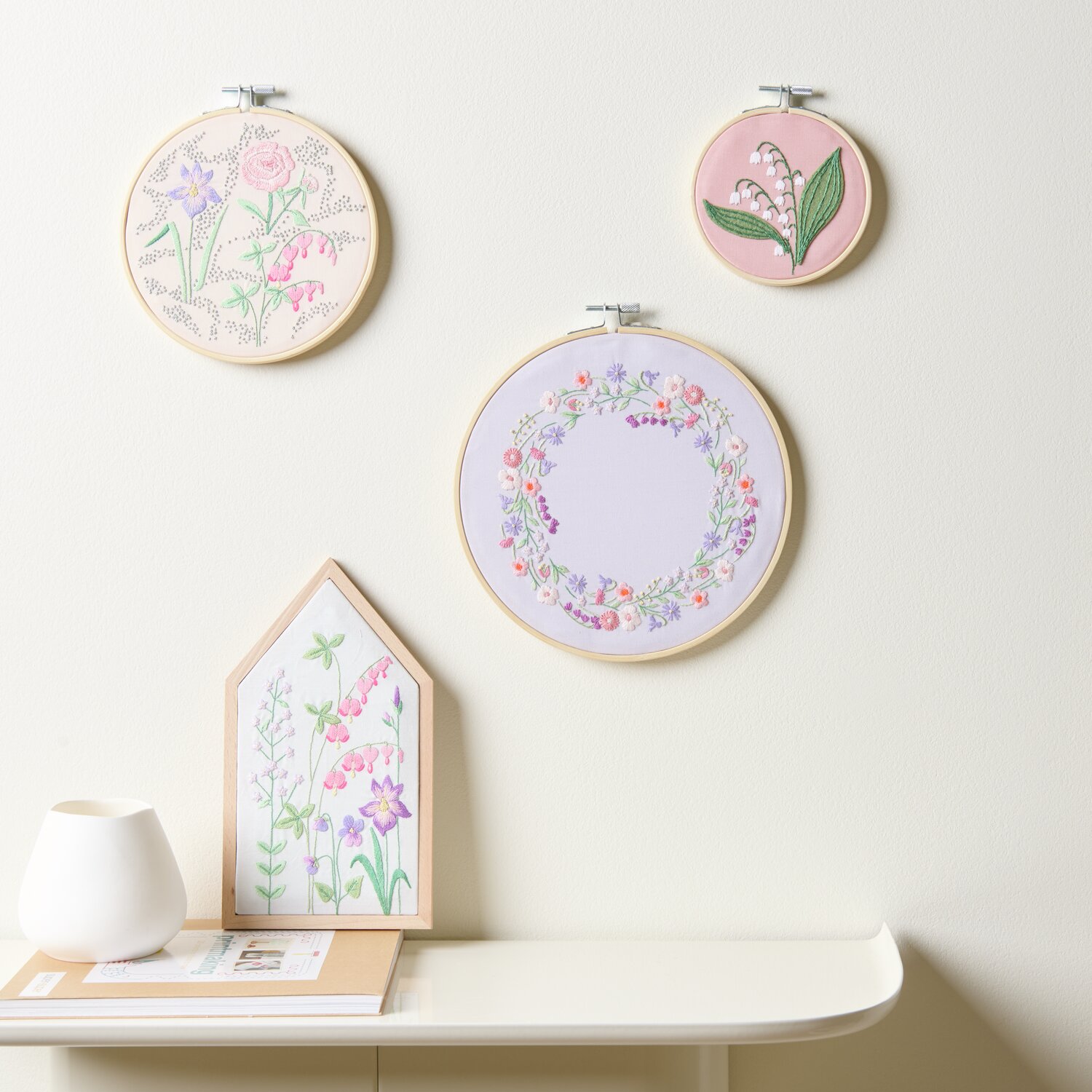 Stickpackung Vorgezeichnet Frühlingshafte Blumenwiese: Drei Stickrahmen mit floralen Motiven in Pastellfarben an einer Wand.