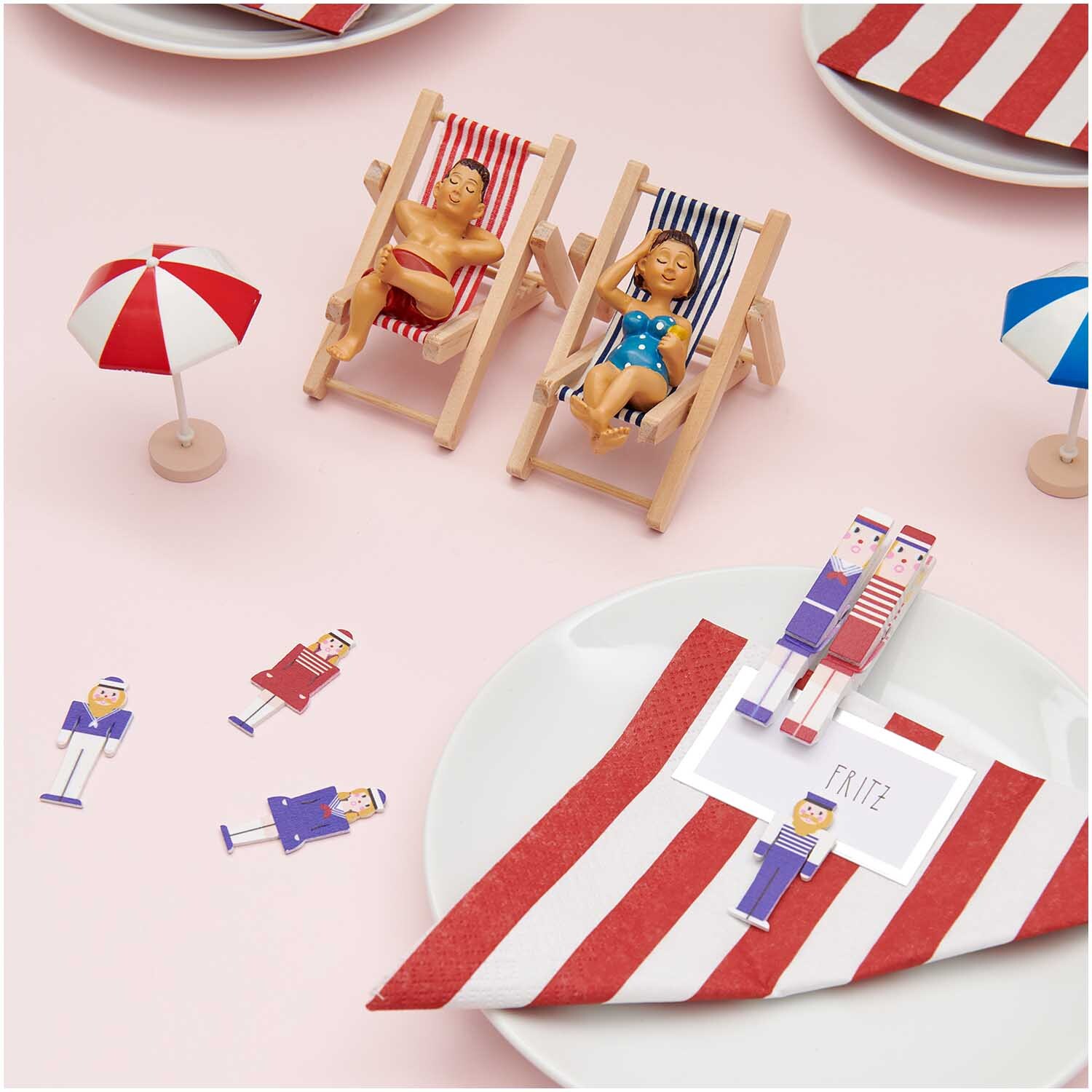 Deko-Liegestuhl rot-weiß von Rico Design: Miniatur-Liegestühle mit Puppen, umgeben von rot-weißen Accessoires auf einem Tisch.