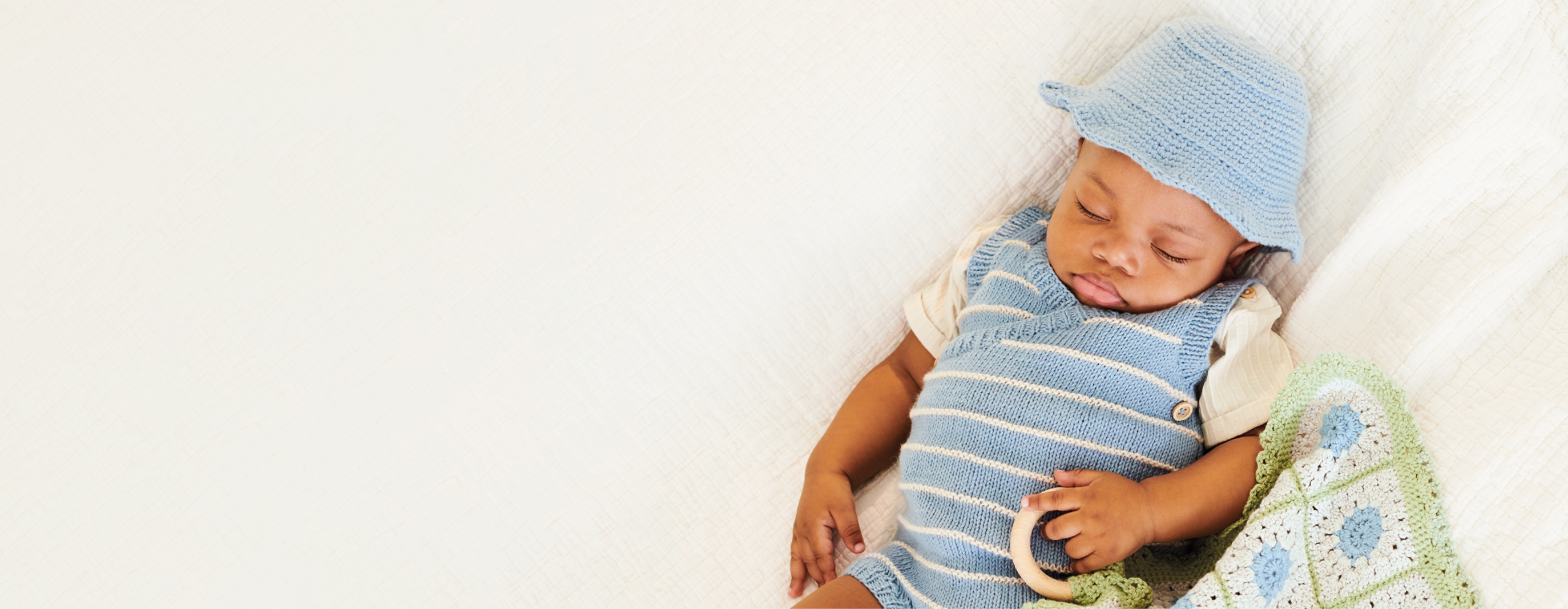 Baby mit gestricktem Outfit und Mütze hält ein gehäkeltes Tuch in der Hand und liegt auf einer Decke.