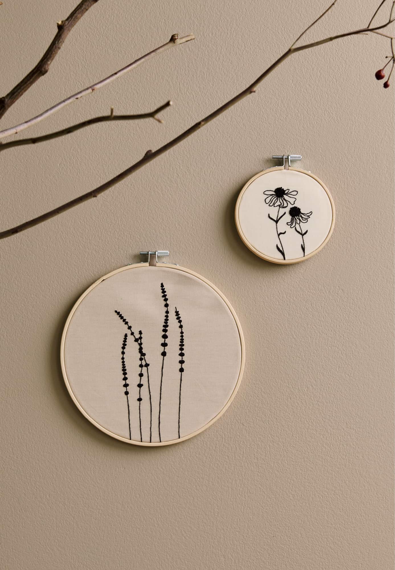 Zwei Stickbilder mit Motiven von Gräsern und Blumen hängen an einer beigen Wand.