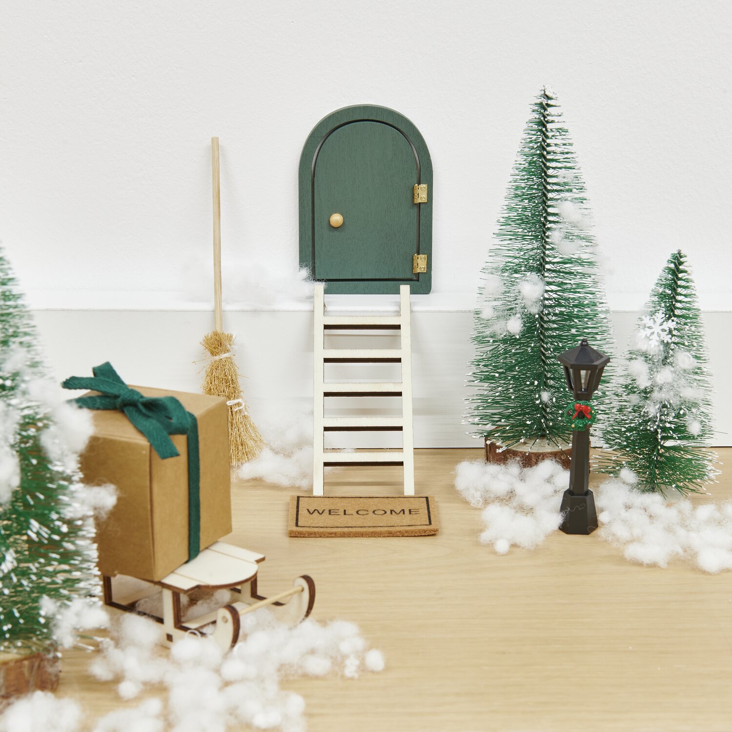 Miniatur Wichteltür rund mit grüner Tür, Leiter und Willkommensschild, umgeben von Tannenbäumen und Dekoration.