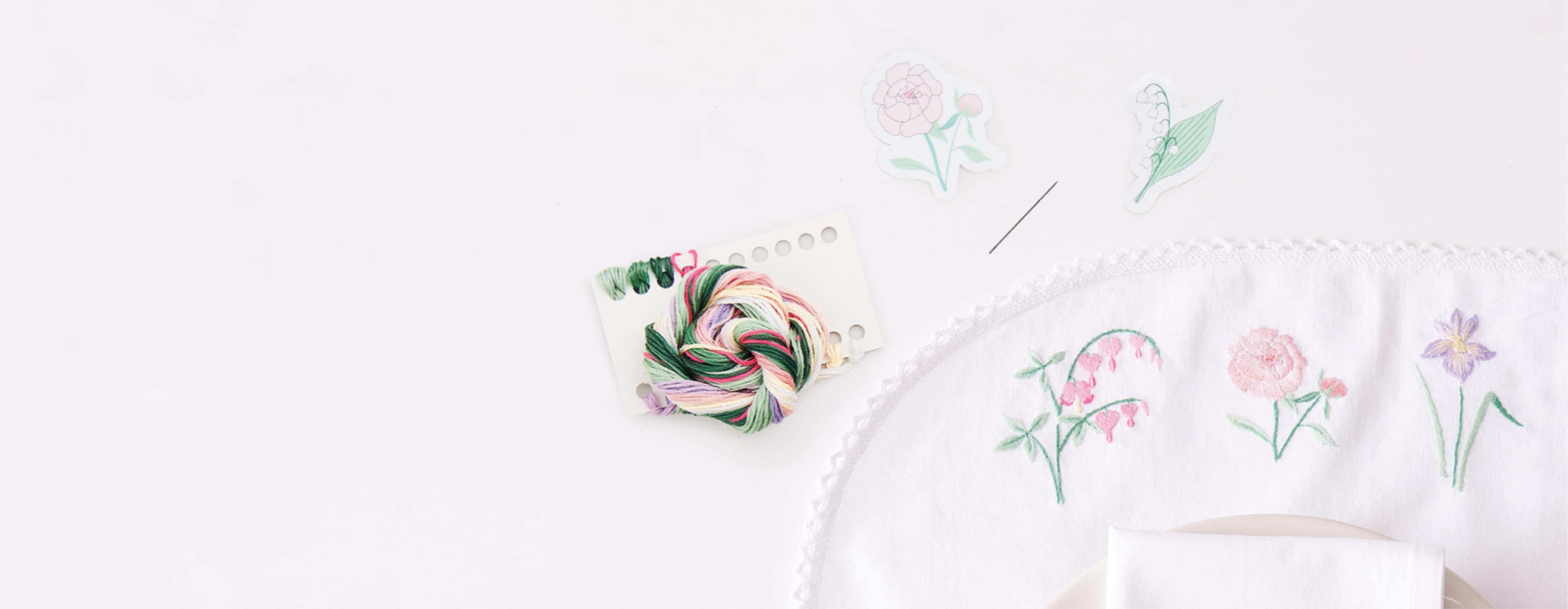Mit Blumen besticktes Tischset liegt umgeben von floralen Stick-and-Stitch-Vorlagen und Stickzubehör auf weißem Hintergrund.