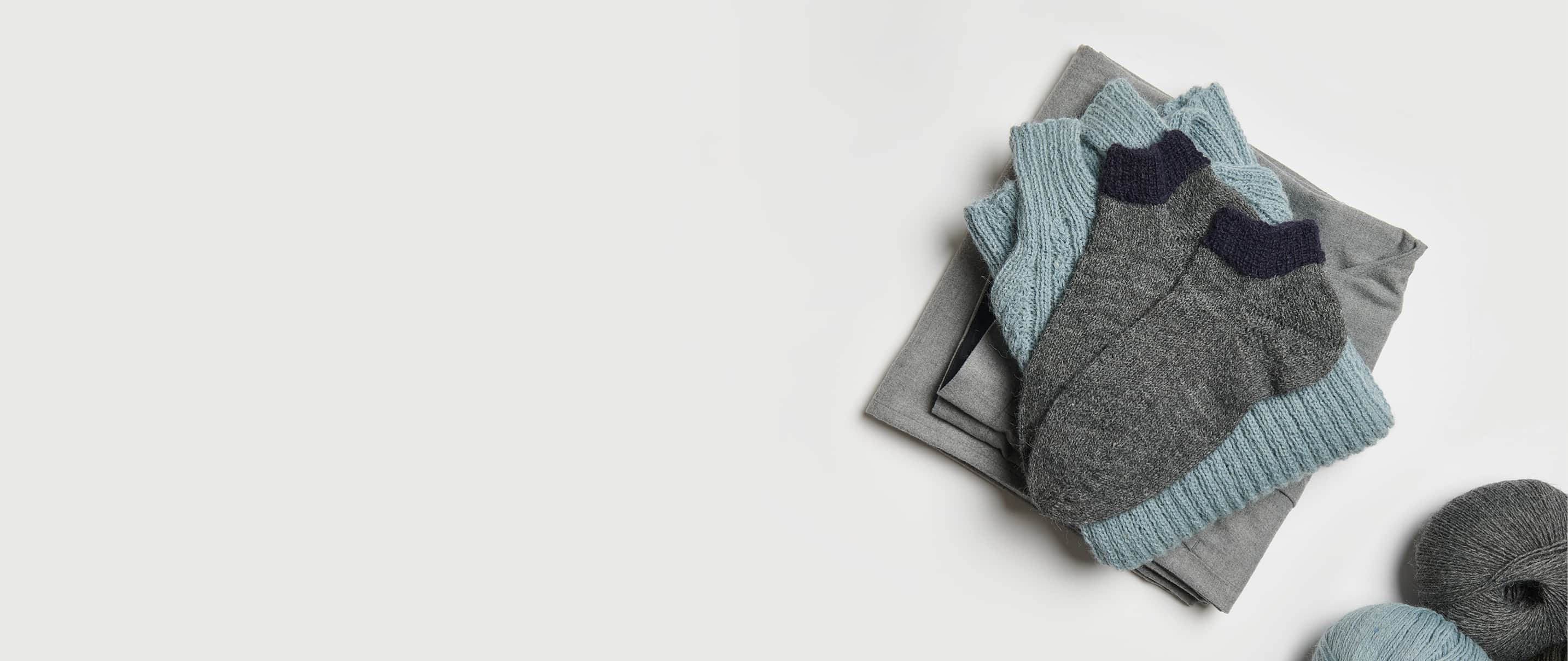 Gestrickte Socken in Grau und Schwarz liegen auf einem Stapel von Kleidungsstücken. Wollknäuel in den Farben liegen daneben.