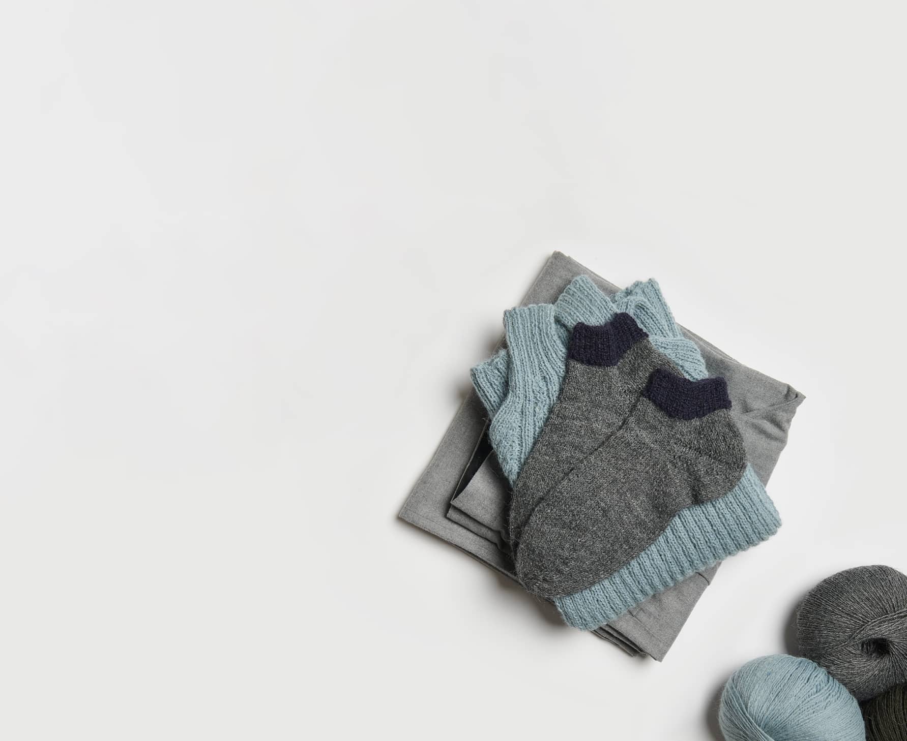 Gestrickte Socken in Grau und Schwarz liegen auf einem Stapel von Kleidungsstücken. Wollknäuel in den Farben liegen daneben.