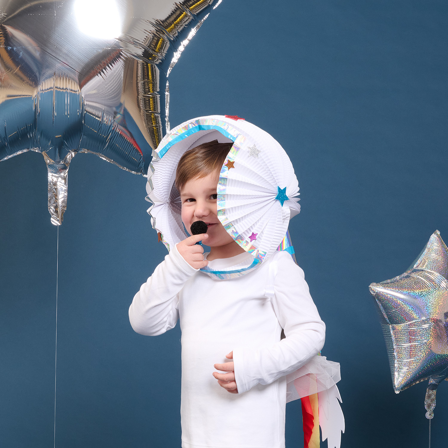 Bastelanleitung Astronauten-Kostüm: Kind in weißem Kostüm mit Helm aus Papier, umgeben von silbernen Sternballons.