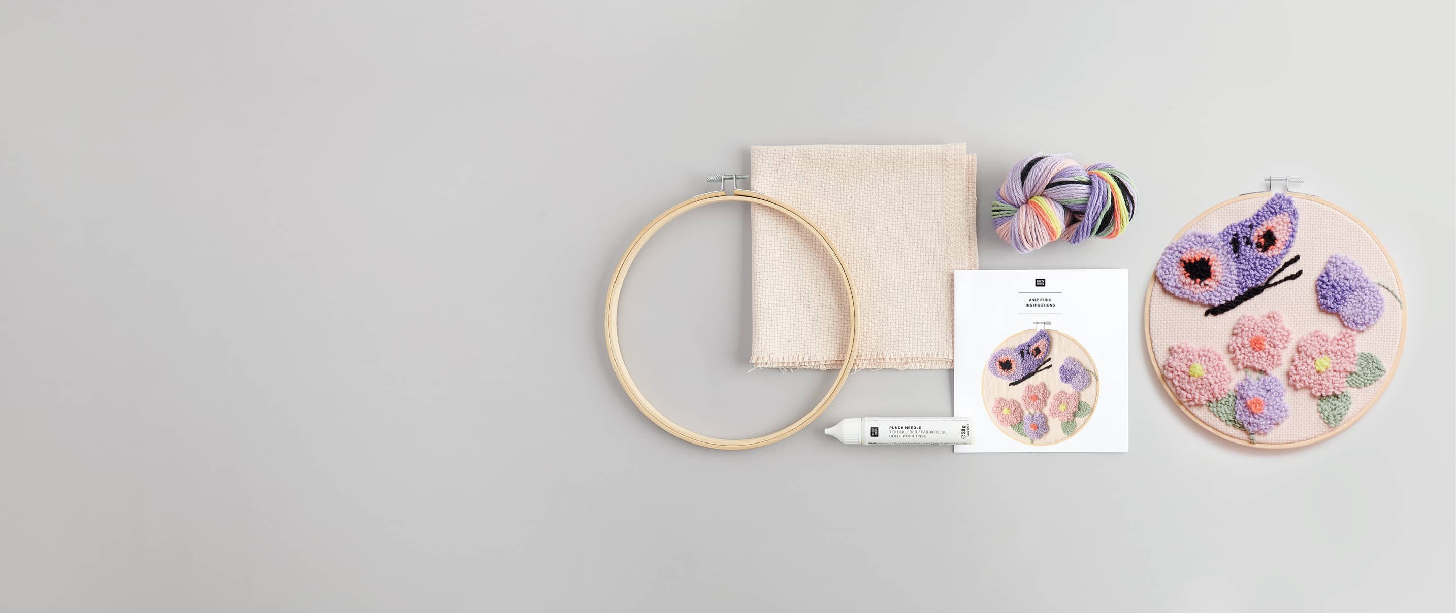 Punch Needle Bild mit Blumen und Schmetterling liegt neben Stickrahmen, Anleitungsbogen, Wolle und Stoff auf grauem Hintergrund angeordnet