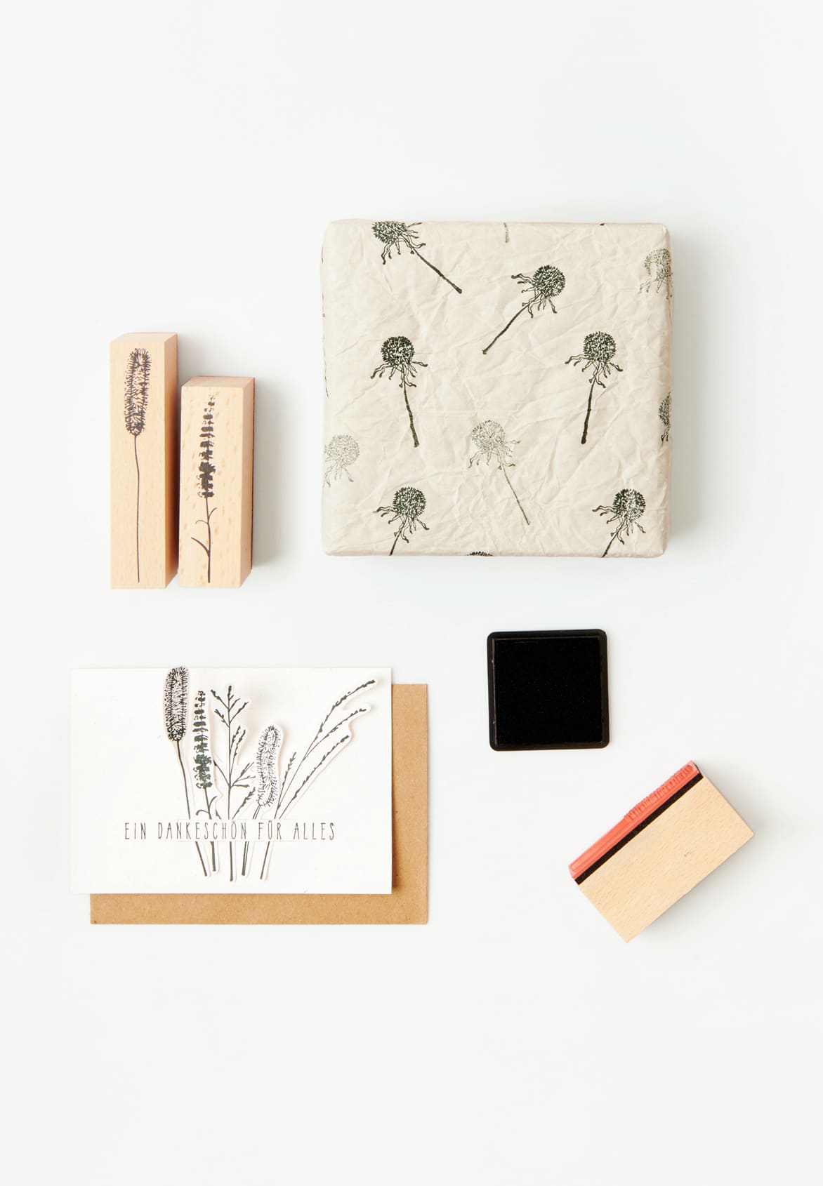 Bestempelte Karte und Geschenk liegen umgeben von Holzstempel mit Gräsermotiven und Stempelkissen auf grauem Hintergrund. 