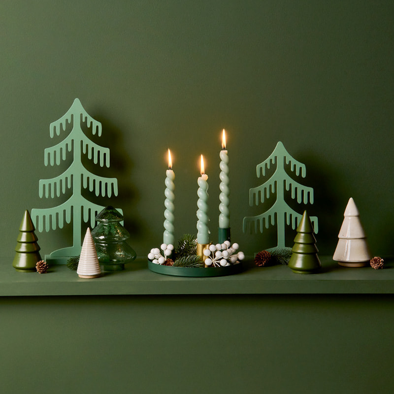 Weihnachtliche Dekofiguren in Form von Tannen und ein geschmücktes Tablett mit Kerzen stehen vor einem dunkelgrünen Hintergrund.