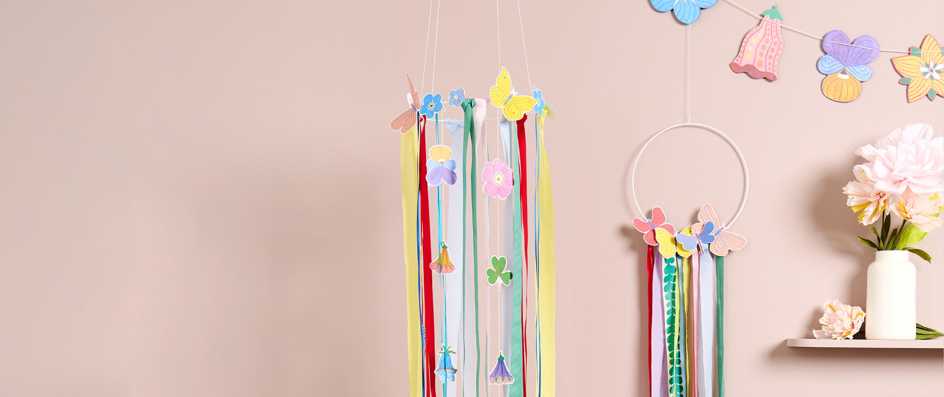 Metallringe sind mit bunten Papierblumen, Schmetterlingen und Bändern zu Kränzen geschmückt und hängen dekorativ vor einer Wand. 