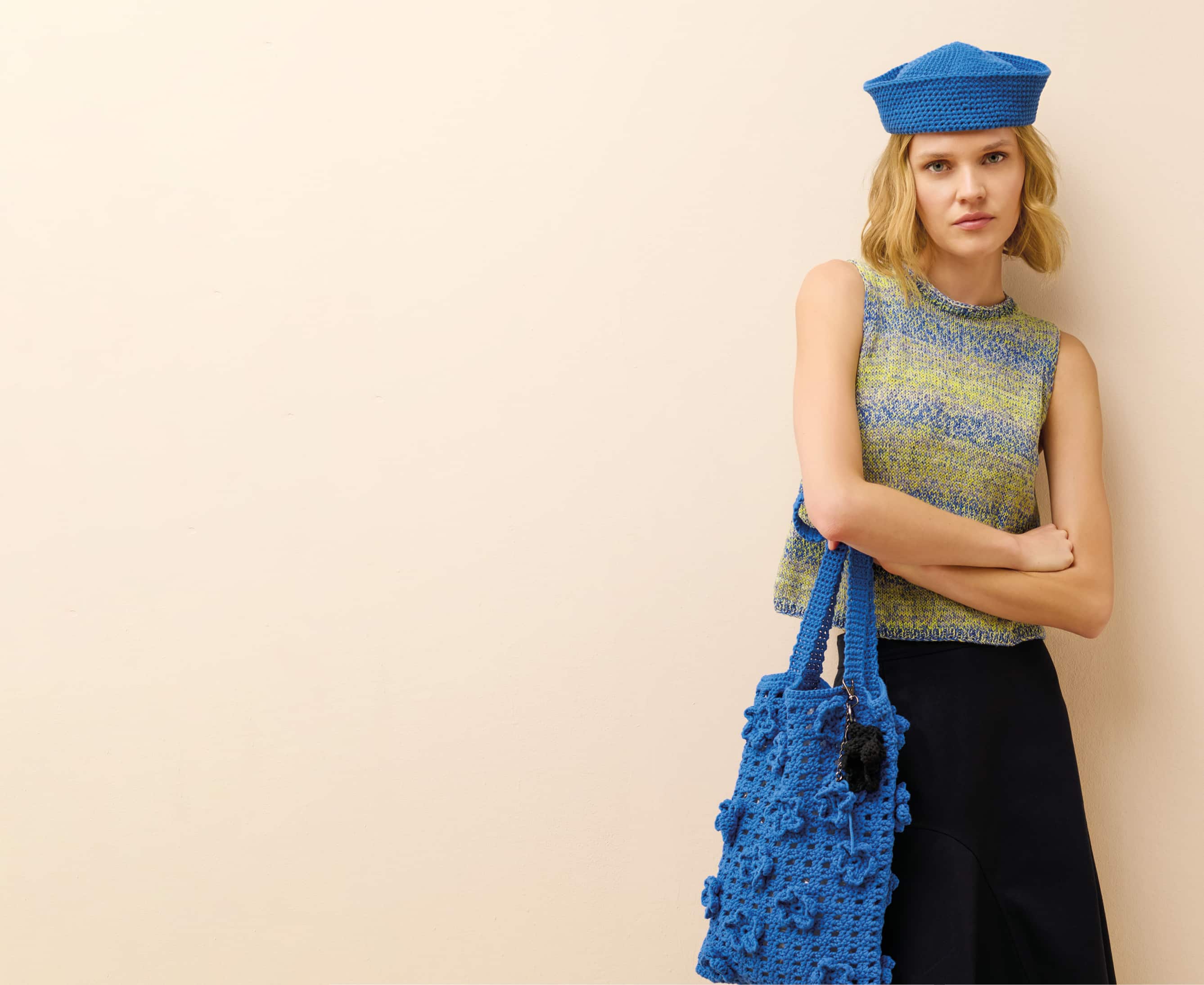 Eine Frau trägt einen gehäkelten Hut, ein gestricktes Top und hält eine gehäkelte Tasche und posiert vor hellem Hintergrund.