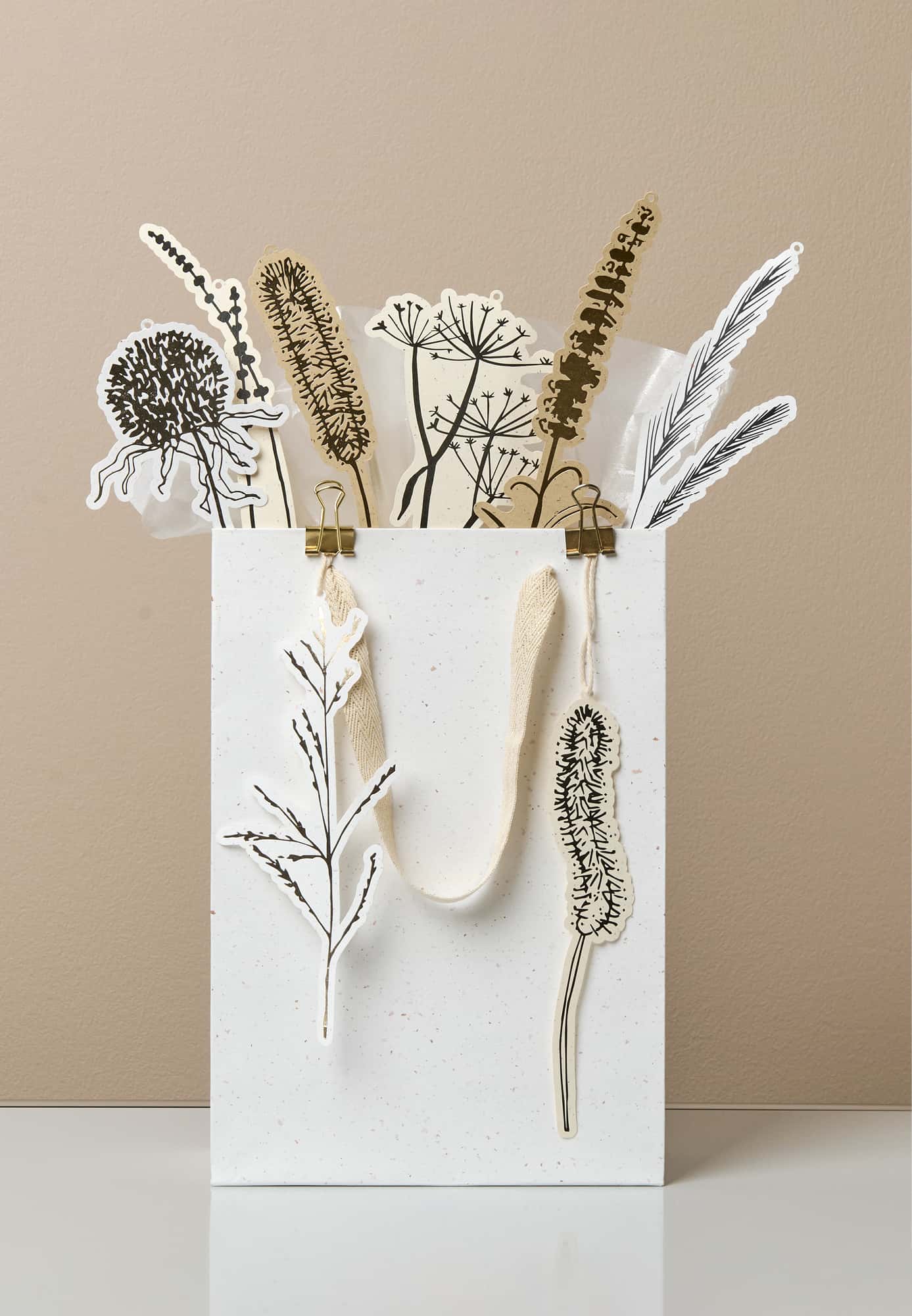 Eine Papiertüte ist mit Papieranhängern mit Trockenblumen-Motiven gefüllt und steht vor beigem Hintergrund. 