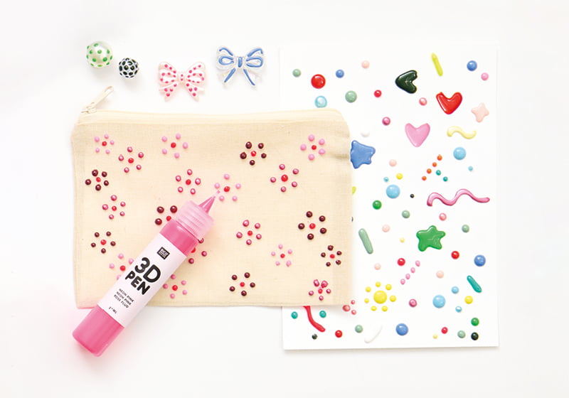 Mit Blumen bemalte Tasche, bunt verzierte Karte und Perlen sind zusammen mit einem 3D Pen in Pink angeordnet.