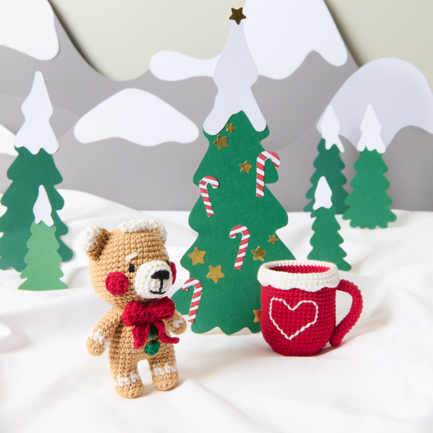 Häkelanleitung Ricorumi CAL Gary the Gingerbread Bear Häkelanleitung Ricorumi CAL Gary the Gingerbread Bear: Ein gehäkelter Bär mit rotem Schal steht neben einer roten Tasse vor winterlicher Kulisse.