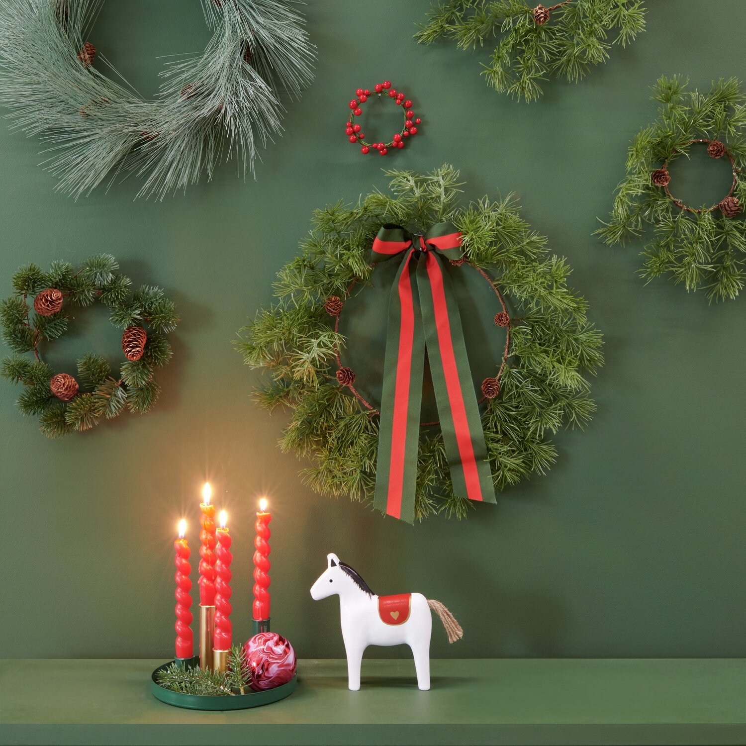 Lärchenkranz mit Zapfen, dekoriert mit roten Bändern, umgeben von weiteren Kränzen an grüner Wand.