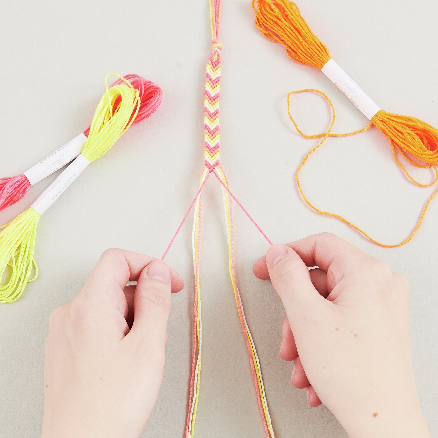 DIY Freundschaftsarmbänder Set Neon Colours: Hände flechten bunte Bänder in Neonfarben.