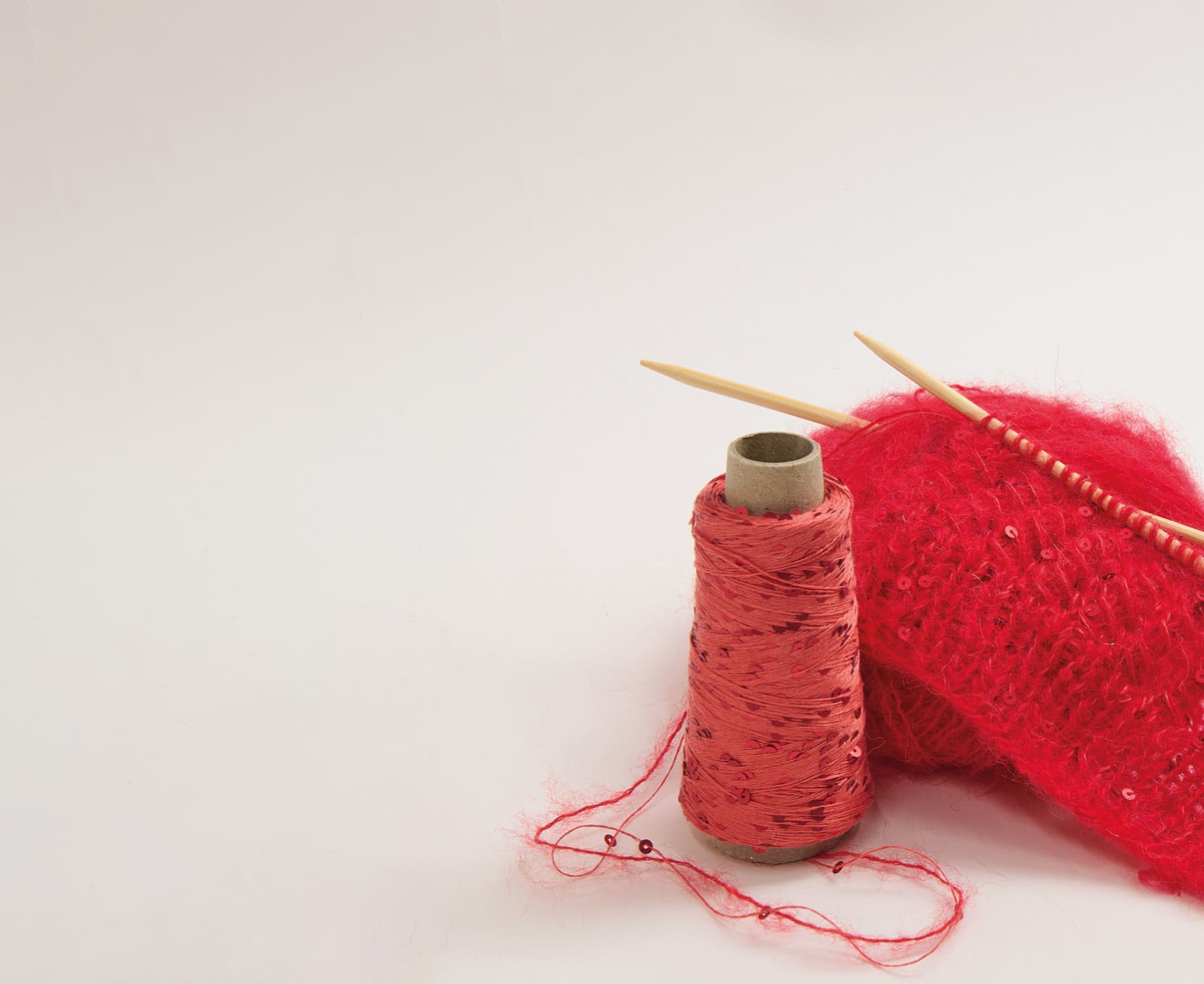 Ein Strickstück auf Stricknadeln steht mit einem roten Paillettengarn und einem roten Garnknäuel vor grauem Hintergrund.