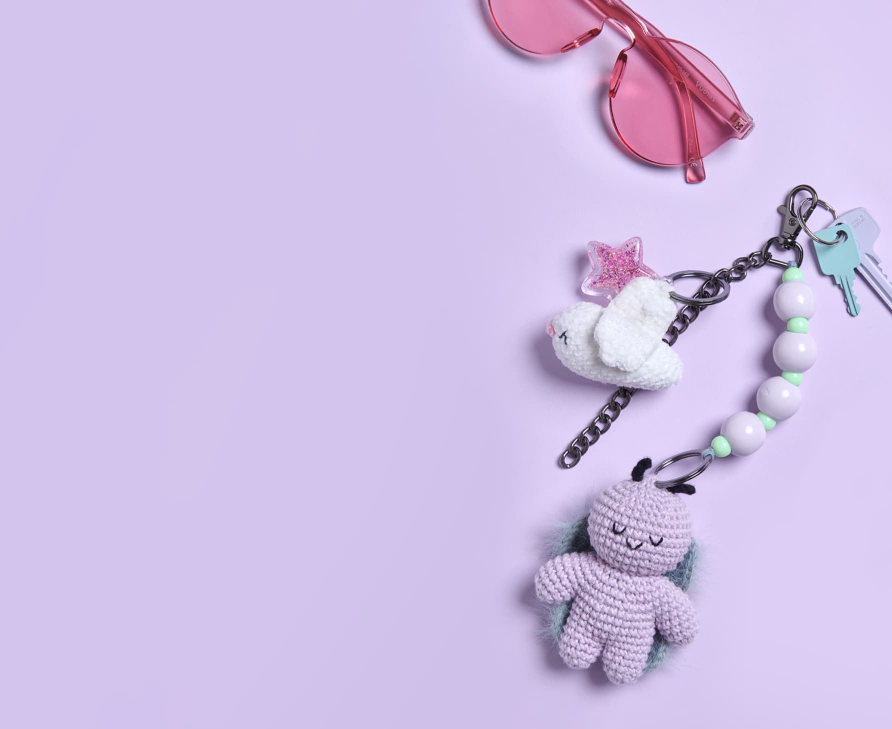 Schlüssel mit gehäkelten Anhängern in Form von einem Vogel und einem Käfer liegen umgeben von einer Sonnenbrille auf lila Hintergrund.