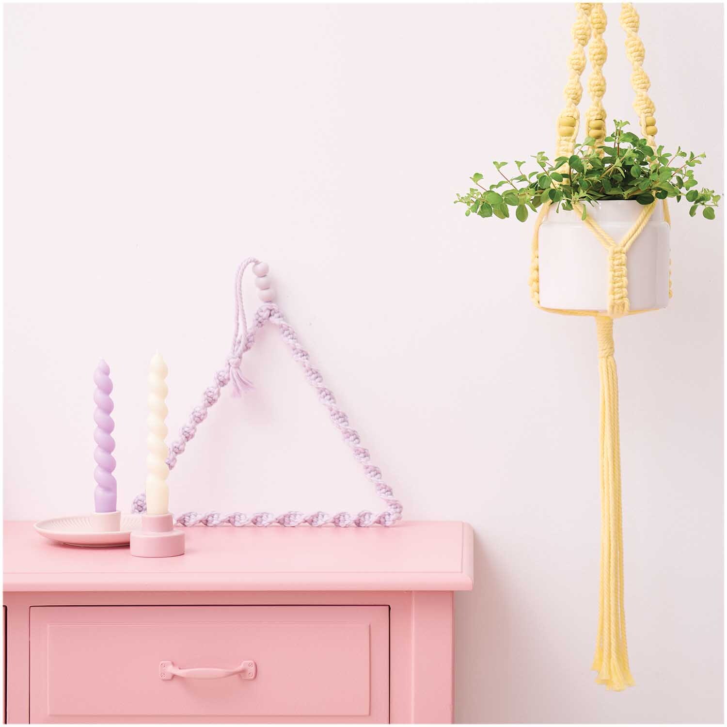 Makramee Set Blumenampel aus Rico Macrame: Gelbe Blumenampel mit Pflanze, dekorativ neben pastellfarbenen Kerzen und rosa Tisch.
