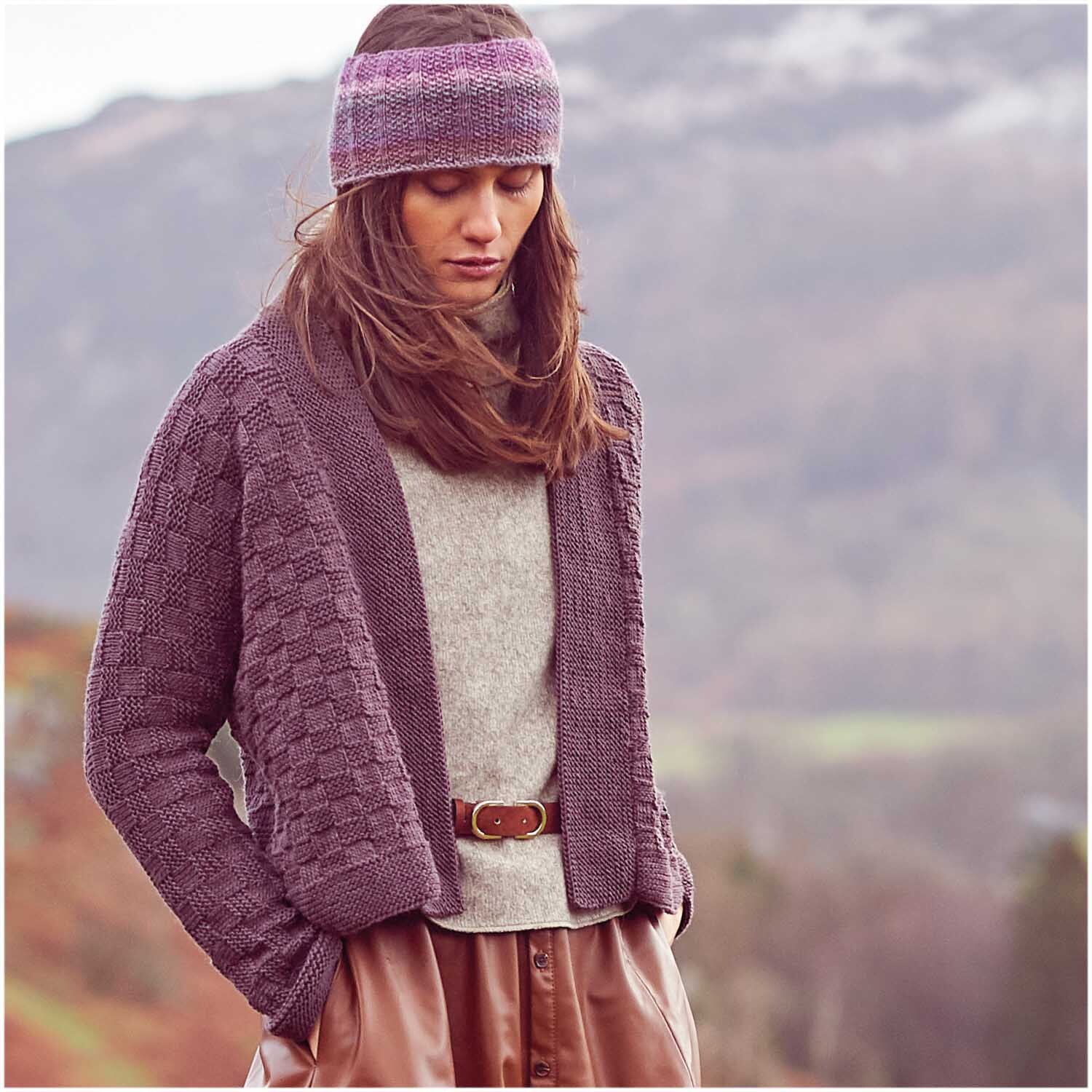 Essentials Merino dk: Frau trägt gestrickte Jacke und Stirnband in lila von Rico Design in Berglandschaft.