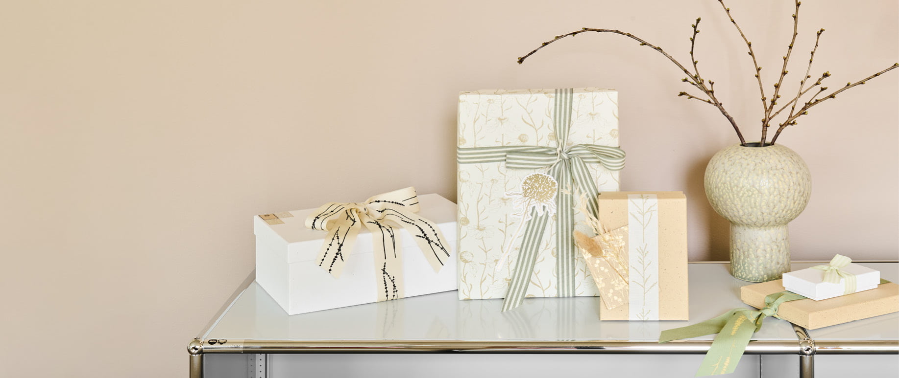 Verpackte Geschenke in Naturtönen und mit Details von Gräsermotiven stehen dekorativ auf einem weißen Regal. 