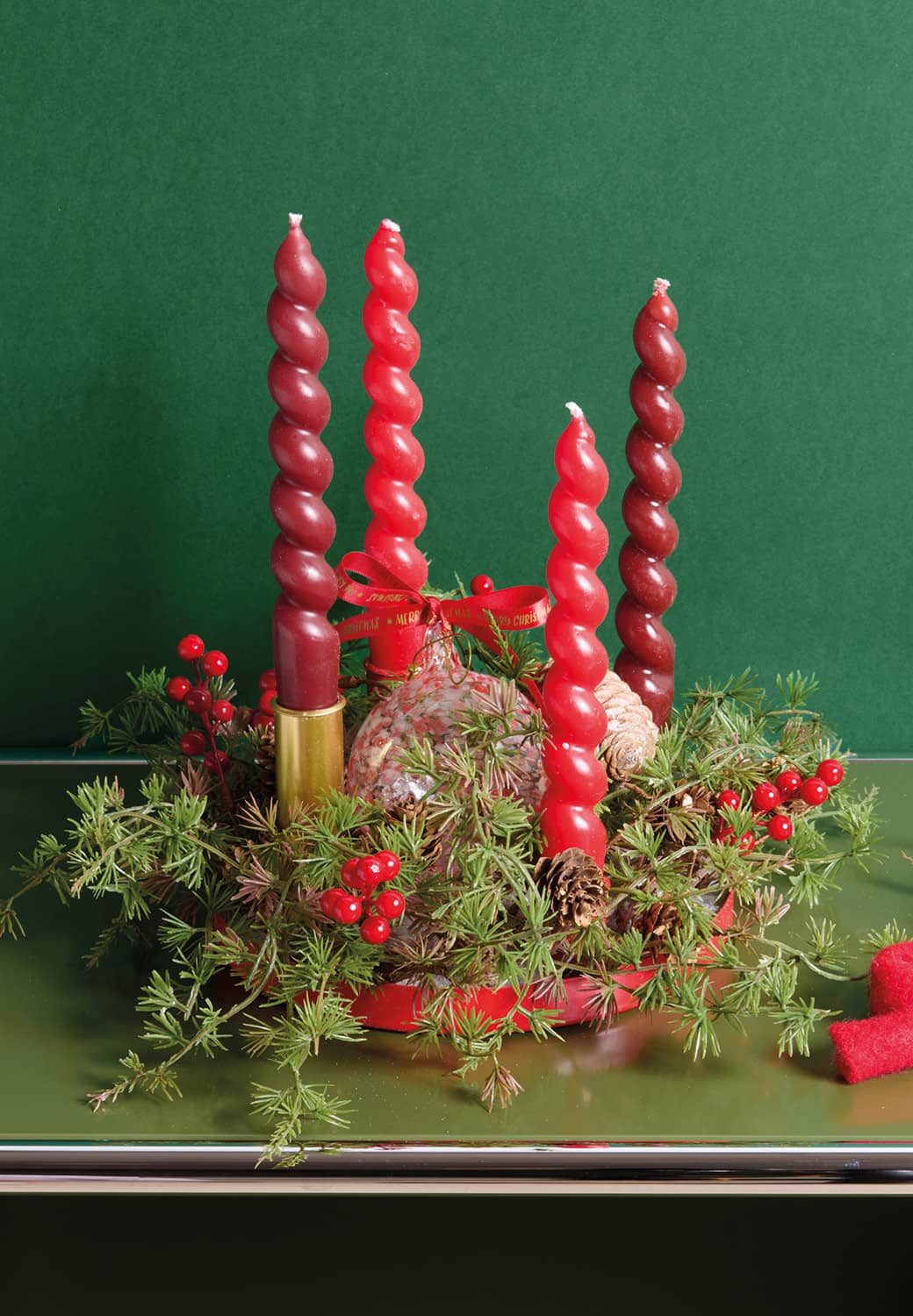 Ein Adventskranz ist vor einer dunkelgrünen Wand mit Tannenzweigen, Beeren und roten Kerzen dekoriert.