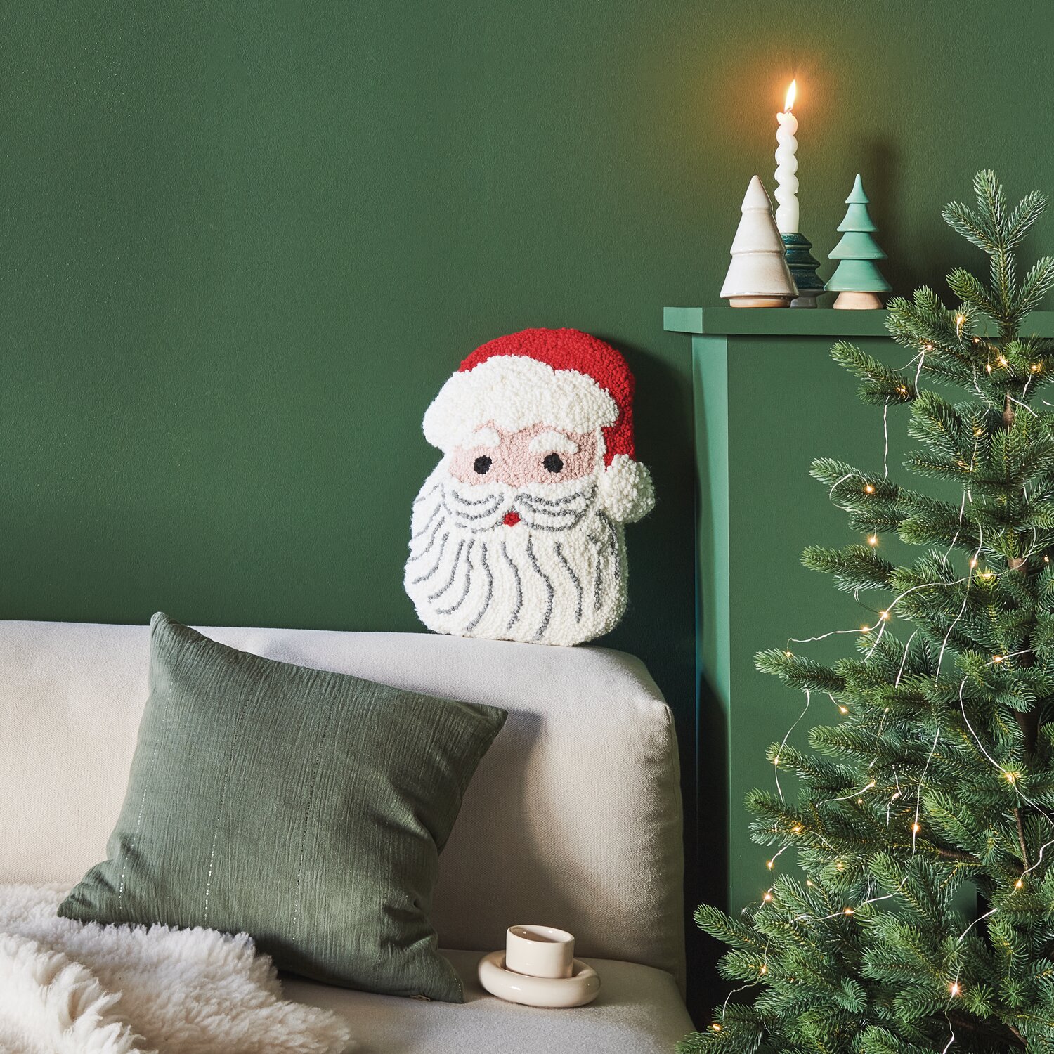 Punch Needle Packung Kissen Santa auf Sofa vor grünem Hintergrund, dekoriert mit Kerzen und Tannenbaum.